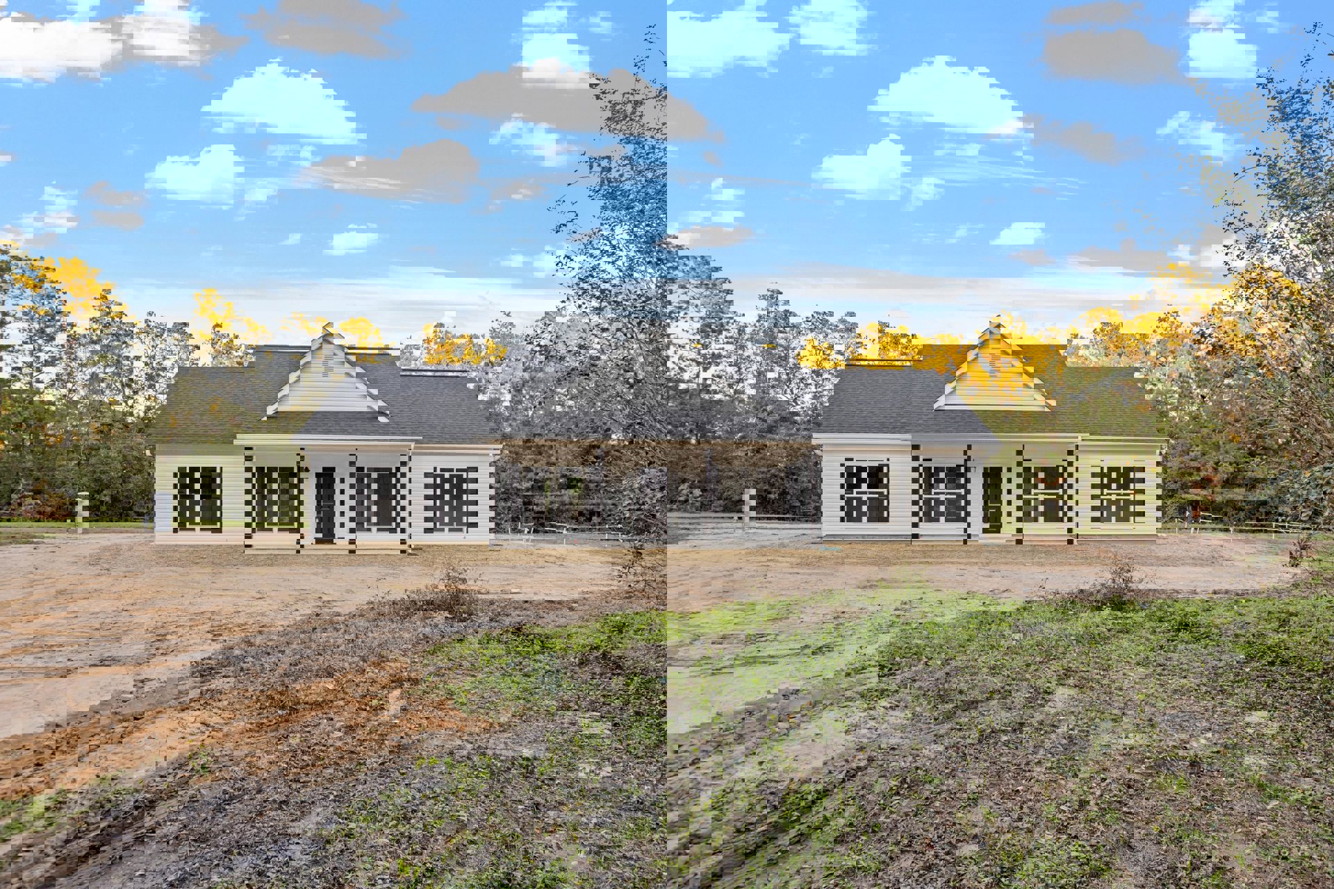 White house with black door and black roof, surrounded by grass, dirt road leading up to the entrance, trees and blue sky with clouds in the background
