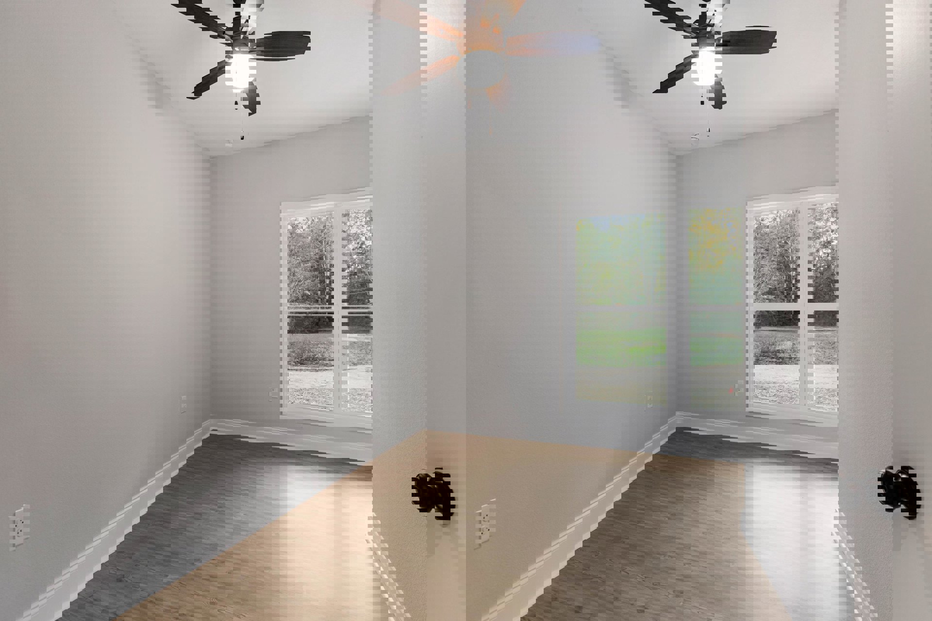 Ceiling fan with light fixture mounted on white plaster ceiling, wood laminate flooring, white electrical outlet on wall, silver doorknob, large window overlooking green trees