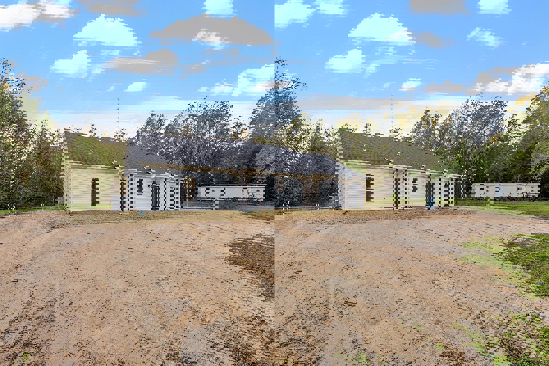 Modern house with black roof, white framed door, surrounded by dirt lot and trees, blue sky with clouds overhead, white trailer parked on grassy area