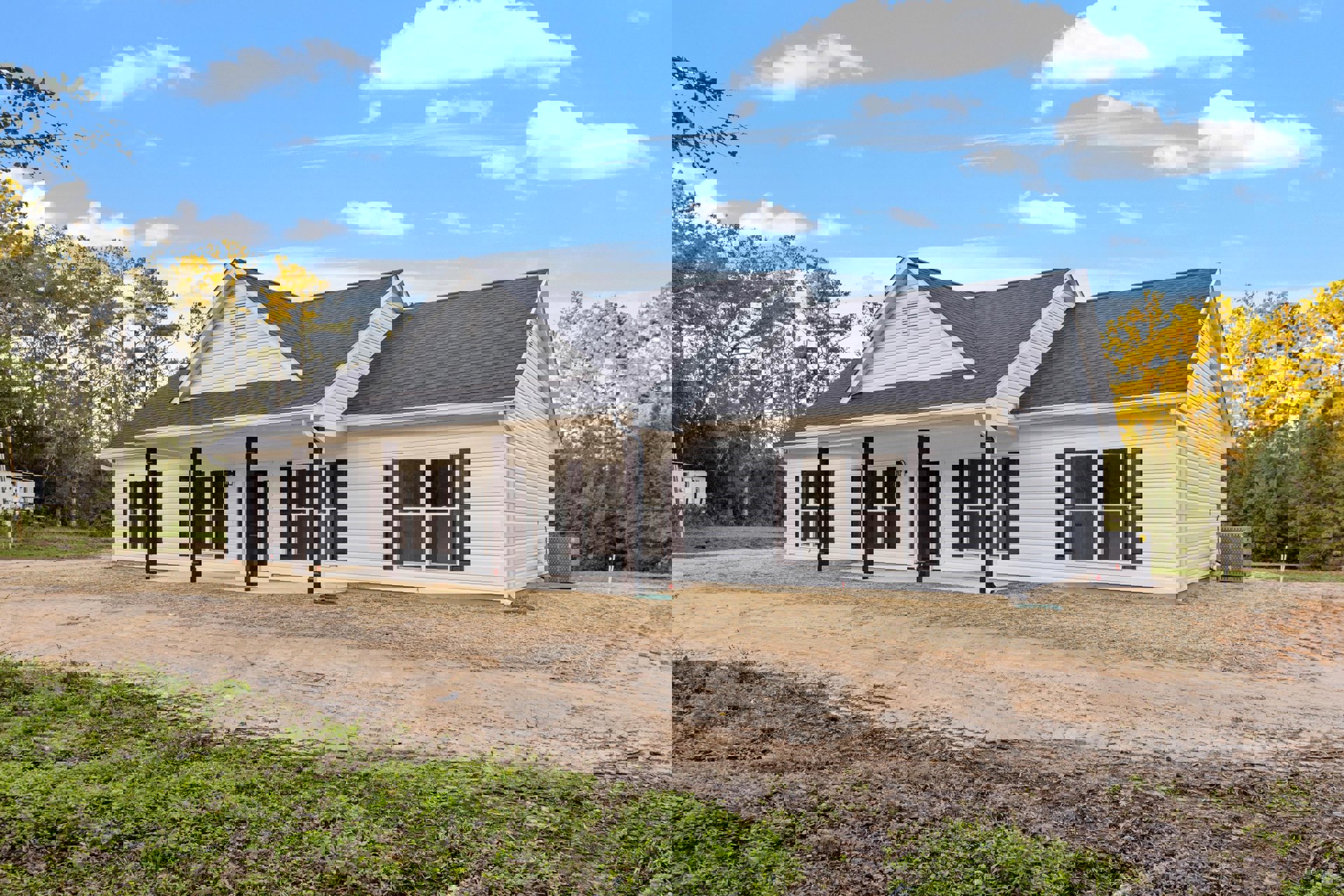 White house with black shutters, dirt road and patch in front, surrounded by trees, blue sky with clouds above, close-up window visible, large grey utility box with holes near