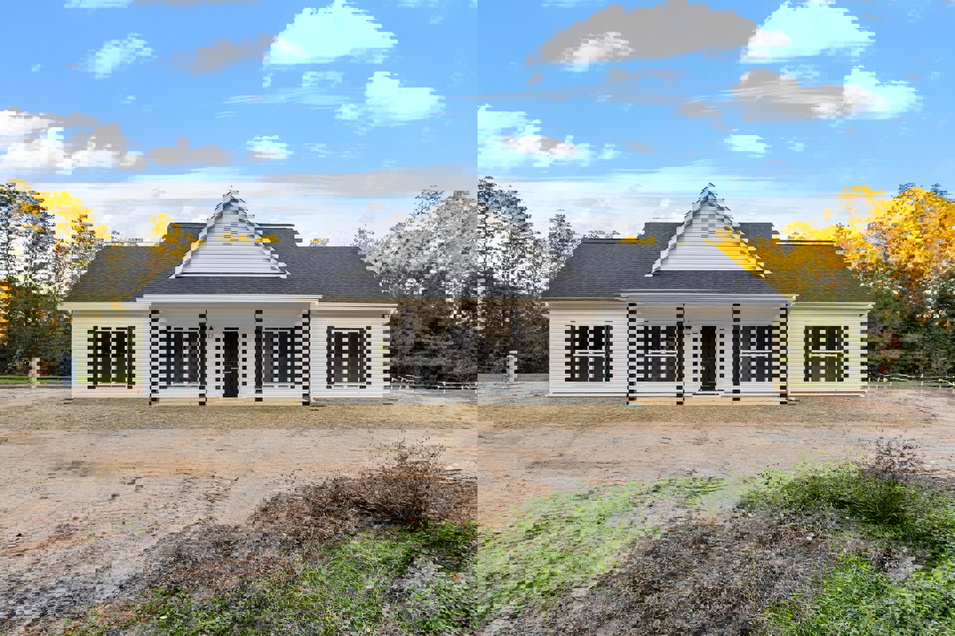 White siding house with black shingle roof, black front door with white trim, dirt driveway, rectangular vent above entry, windows, porch, blue sky with scattered clouds.