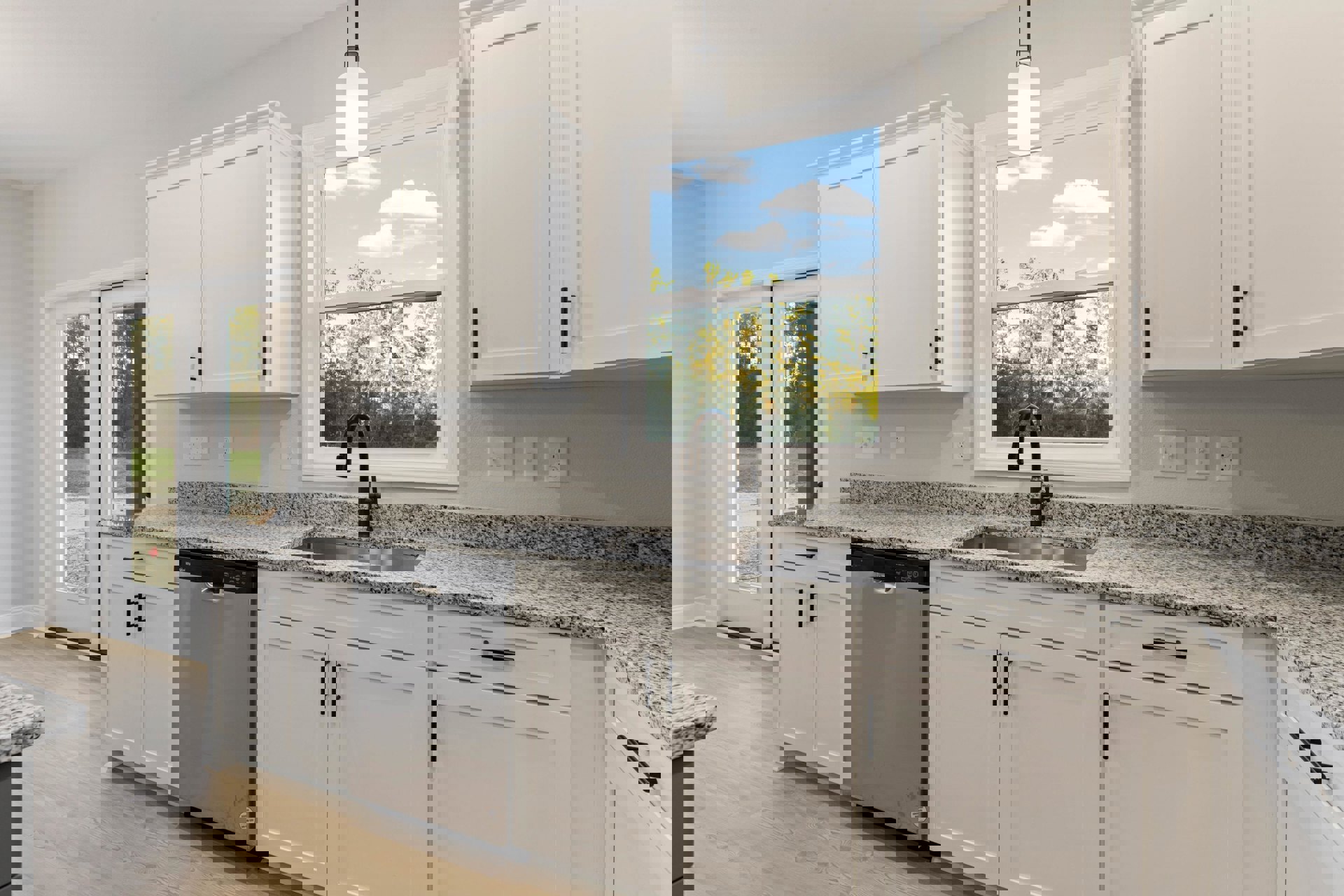 White kitchen with granite countertops, silver refrigerator, black and white speckled sink, white cabinets, black-topped light fixture, window overlooking trees and blue sky