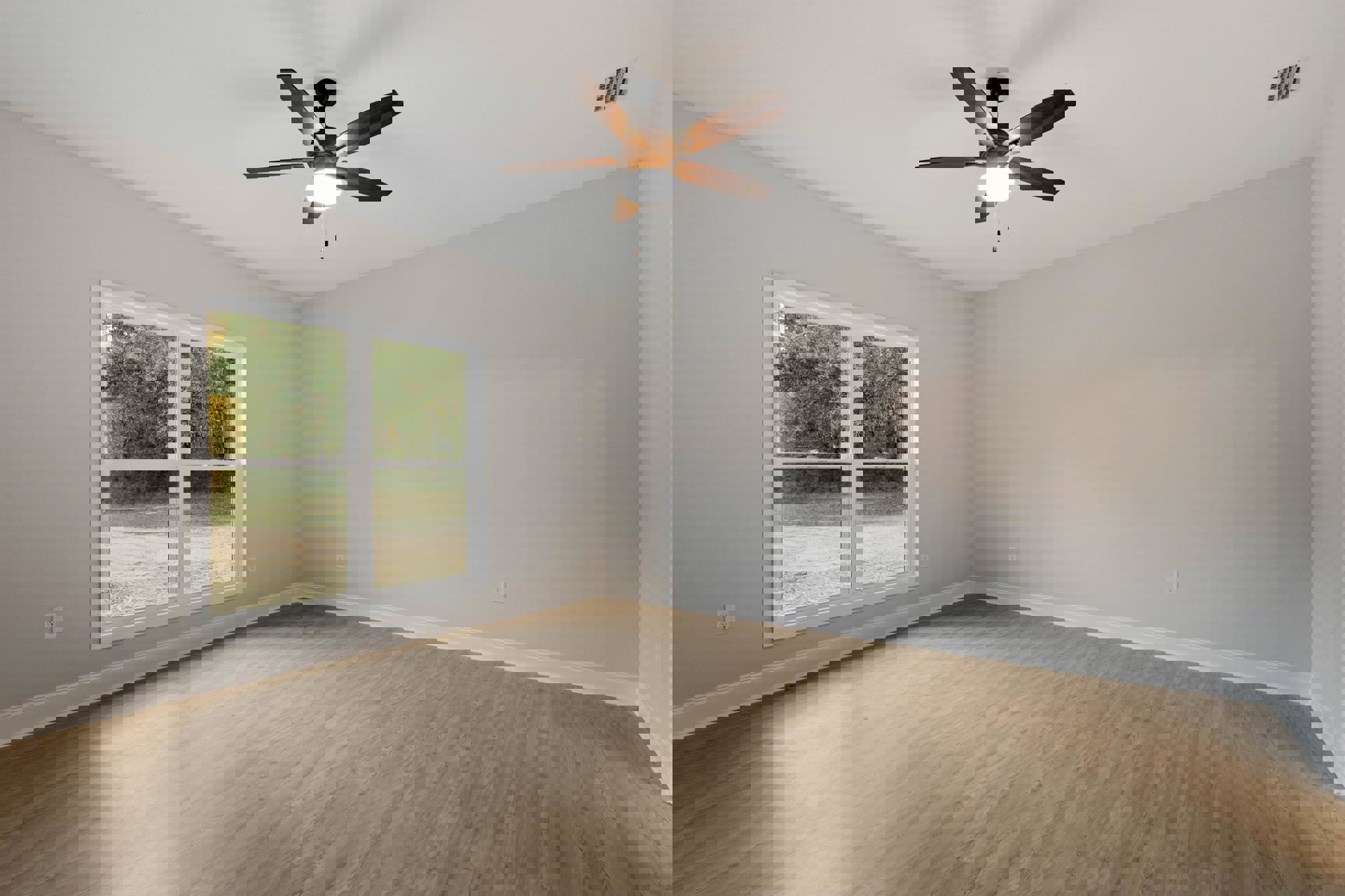 Wood flooring and white walls in a room with a ceiling fan and light fixture, large window overlooking trees, ceiling vent visible