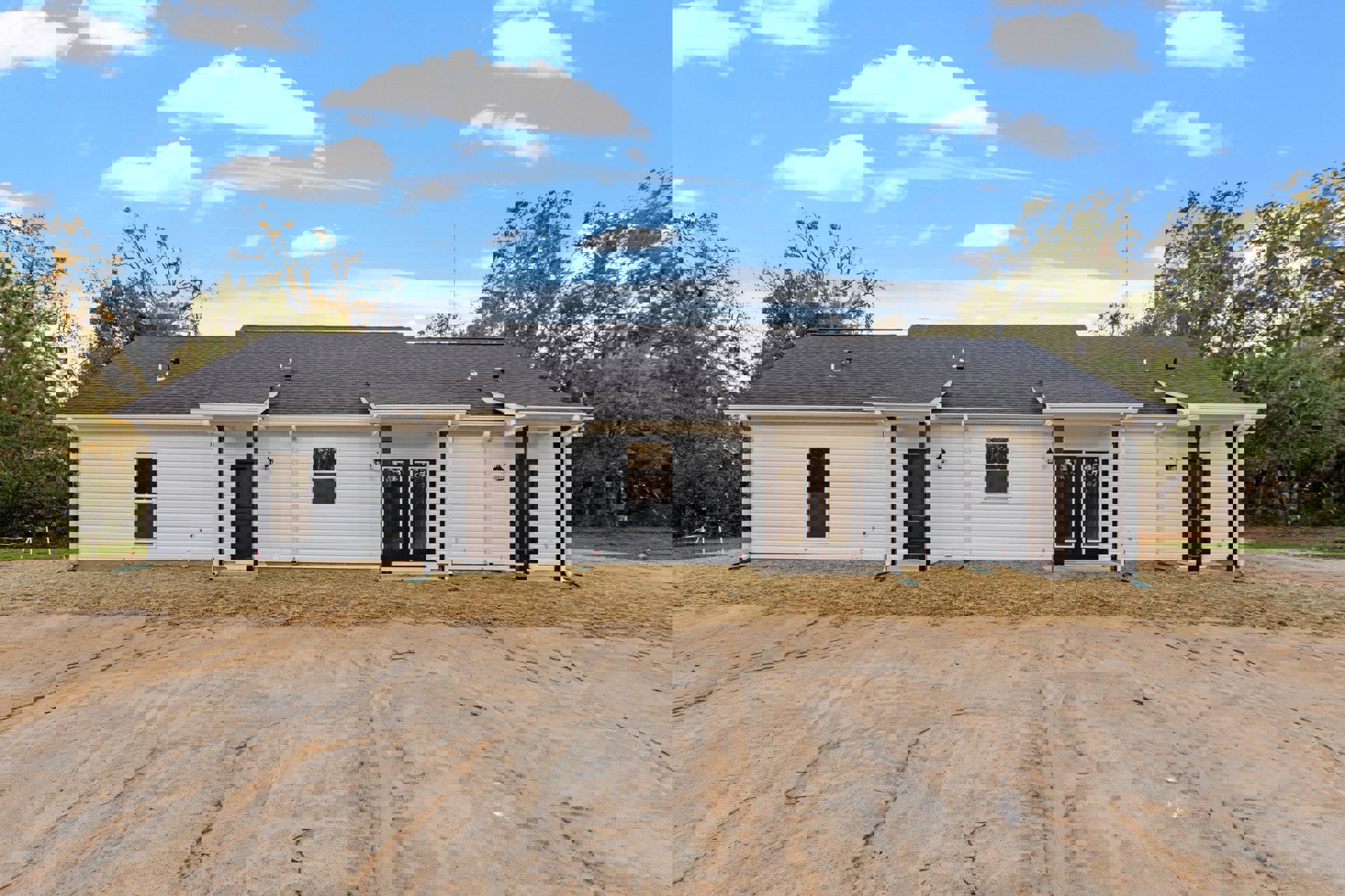 White house with black roof and double doors, set beside a dirt road under a blue sky with scattered clouds; white garage and windows visible, surrounded by open ground and sparse