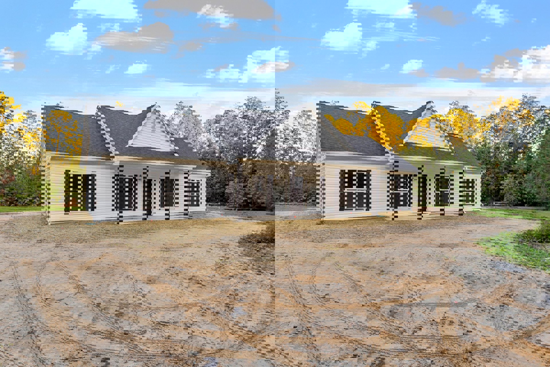 White house with black pillars, tire tracks in sandy dirt yard, white-framed window, blue sky with scattered clouds, sparse trees and plants around property