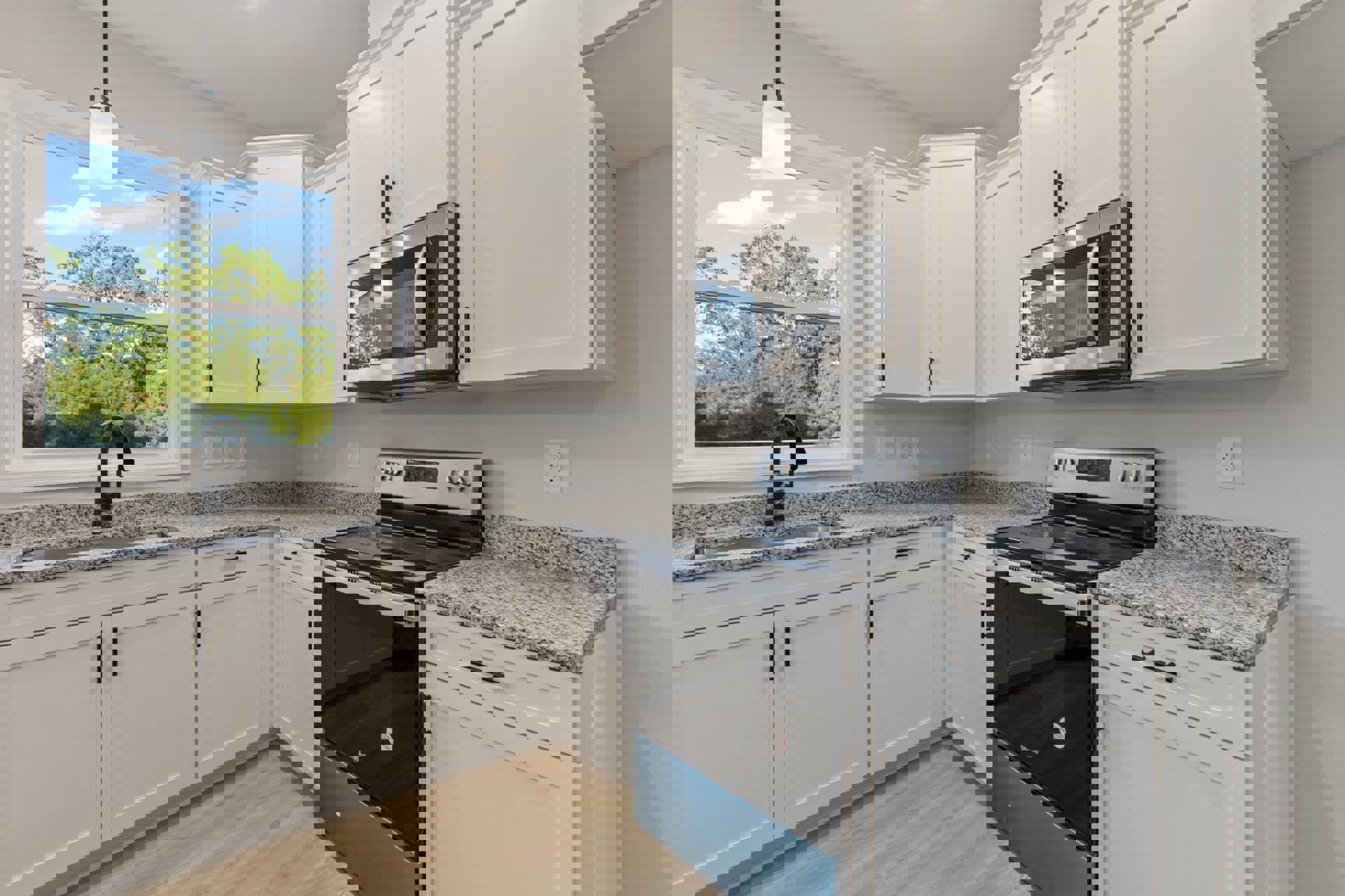 White shaker cabinets, black freestanding stove, stainless microwave with glass door, undermount sink beneath window overlooking trees, quartz countertops, brushed nickel faucet