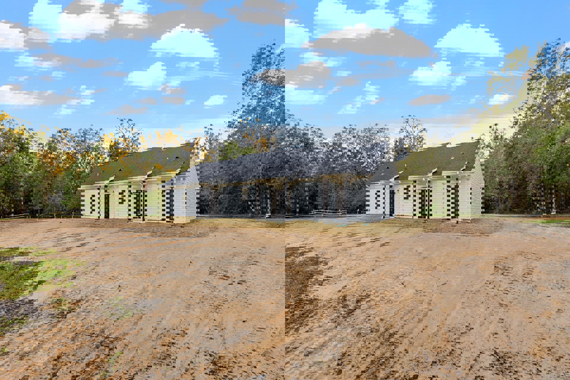 White house with black roof, surrounded by dirt lot, trees in background, and blue sky with scattered clouds