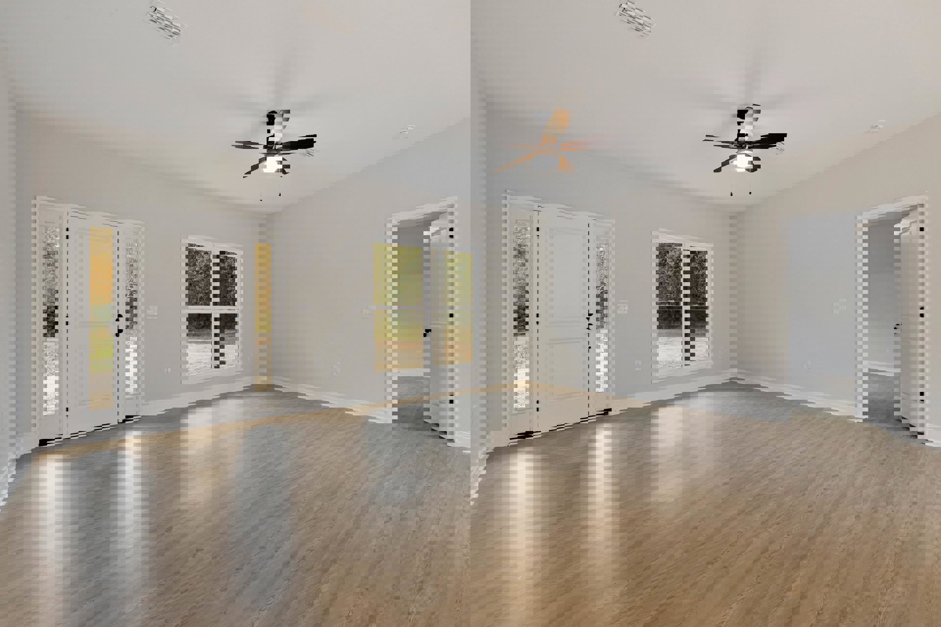 Ceiling fan with light fixture above wood flooring, white door with black knobs and glass panels, large window offering outdoor view, plaster walls and ceiling.