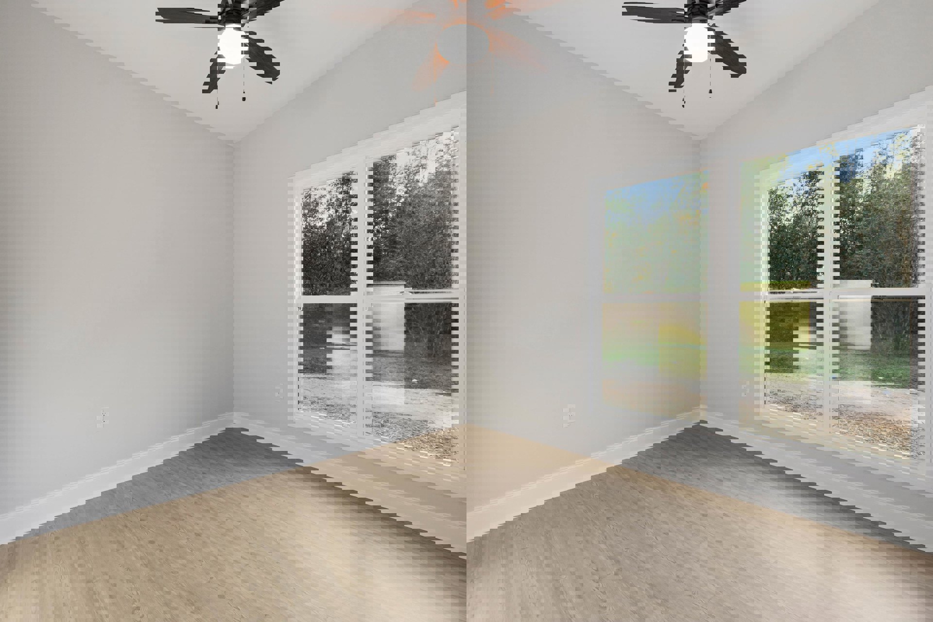 Ceiling fan with light fixture above wood flooring, large window with view of trees, neutral walls and trim