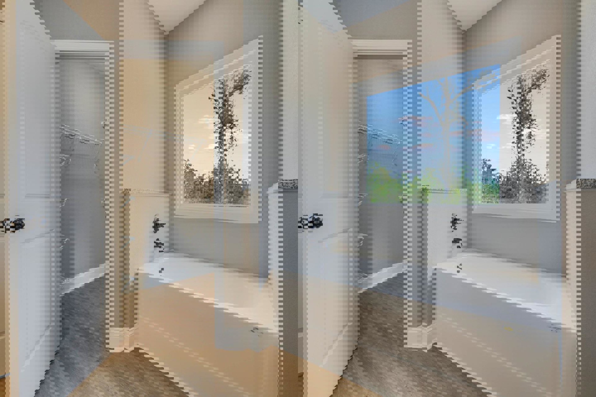 Bathroom with white freestanding tub, glass-enclosed shower, large window overlooking tree, light tile flooring, black spiral towel holder, white metal wall shelf