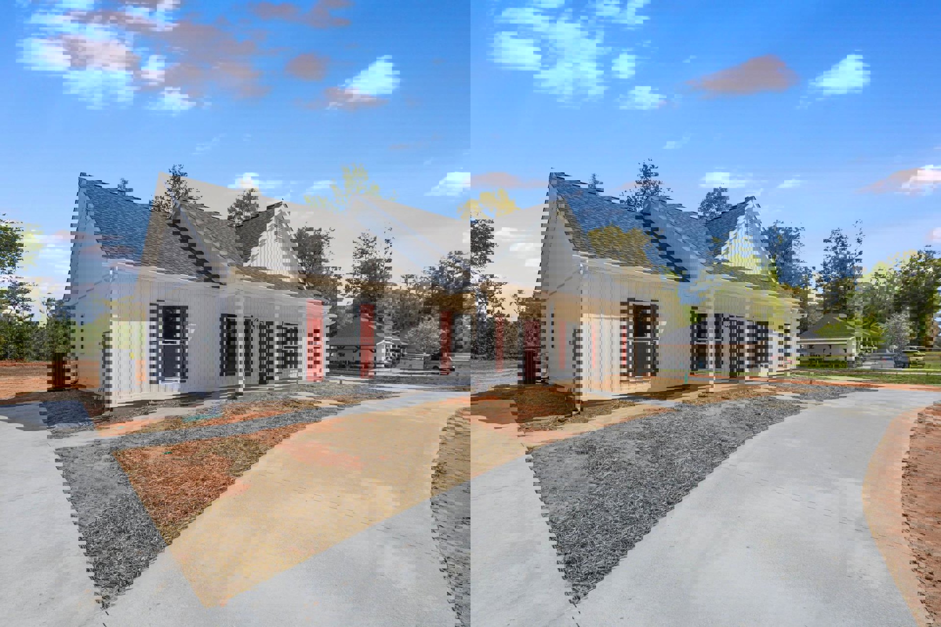 White two-story house with red shutters, white-framed windows, gray shingle roof, concrete driveway bordered by green grass, mature trees behind the home, blue sky with scattered