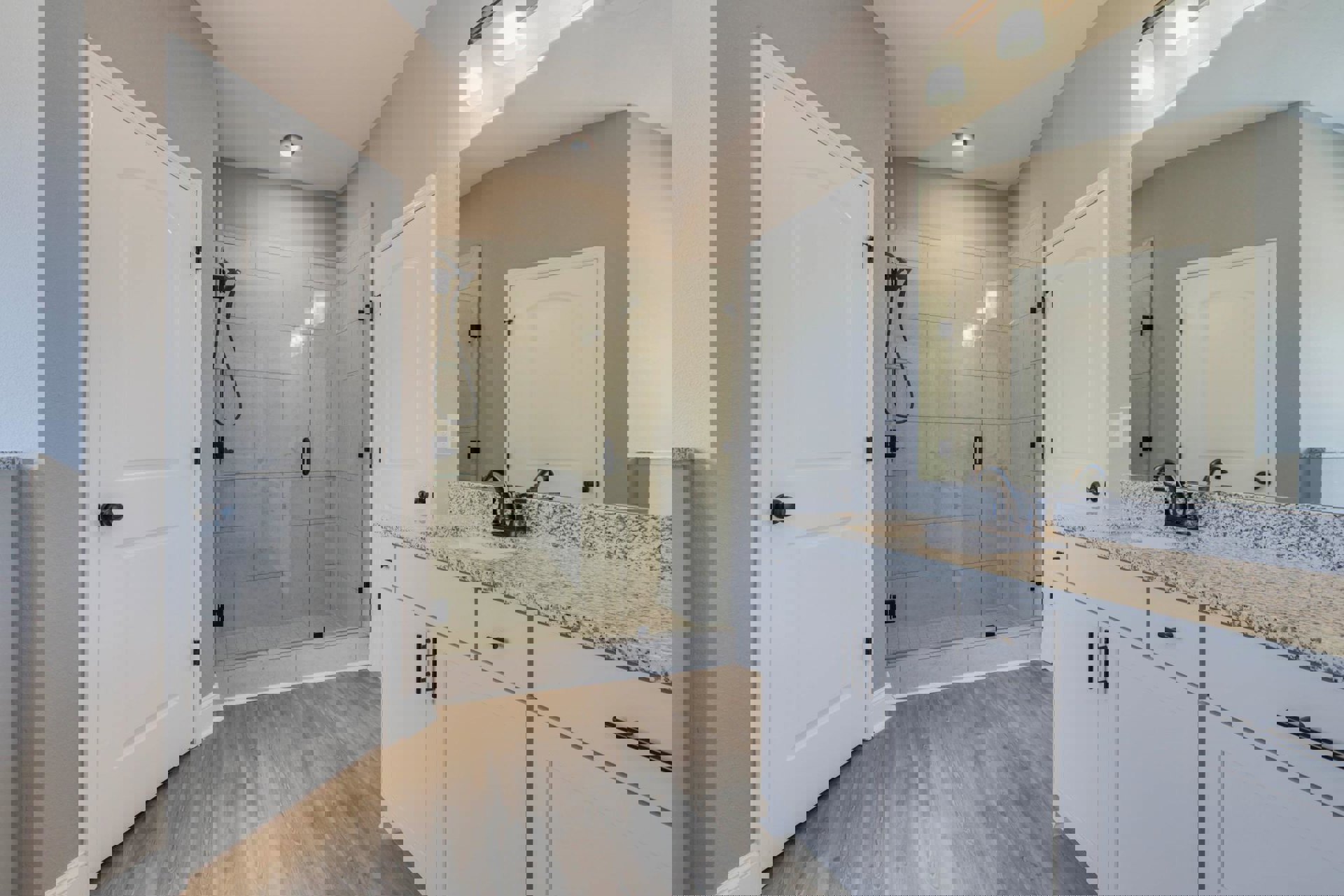 Bathroom with glass shower enclosure, white sink on stone countertop, white door with black knob, tile walls, black cord, and white light fixture with shade