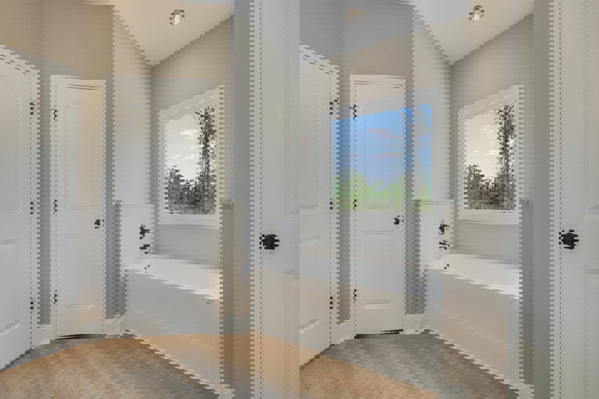 Bathroom with freestanding white bathtub featuring black faucet, white door with black knob, window showing trees outside, light fixture in ceiling, neutral walls and flooring