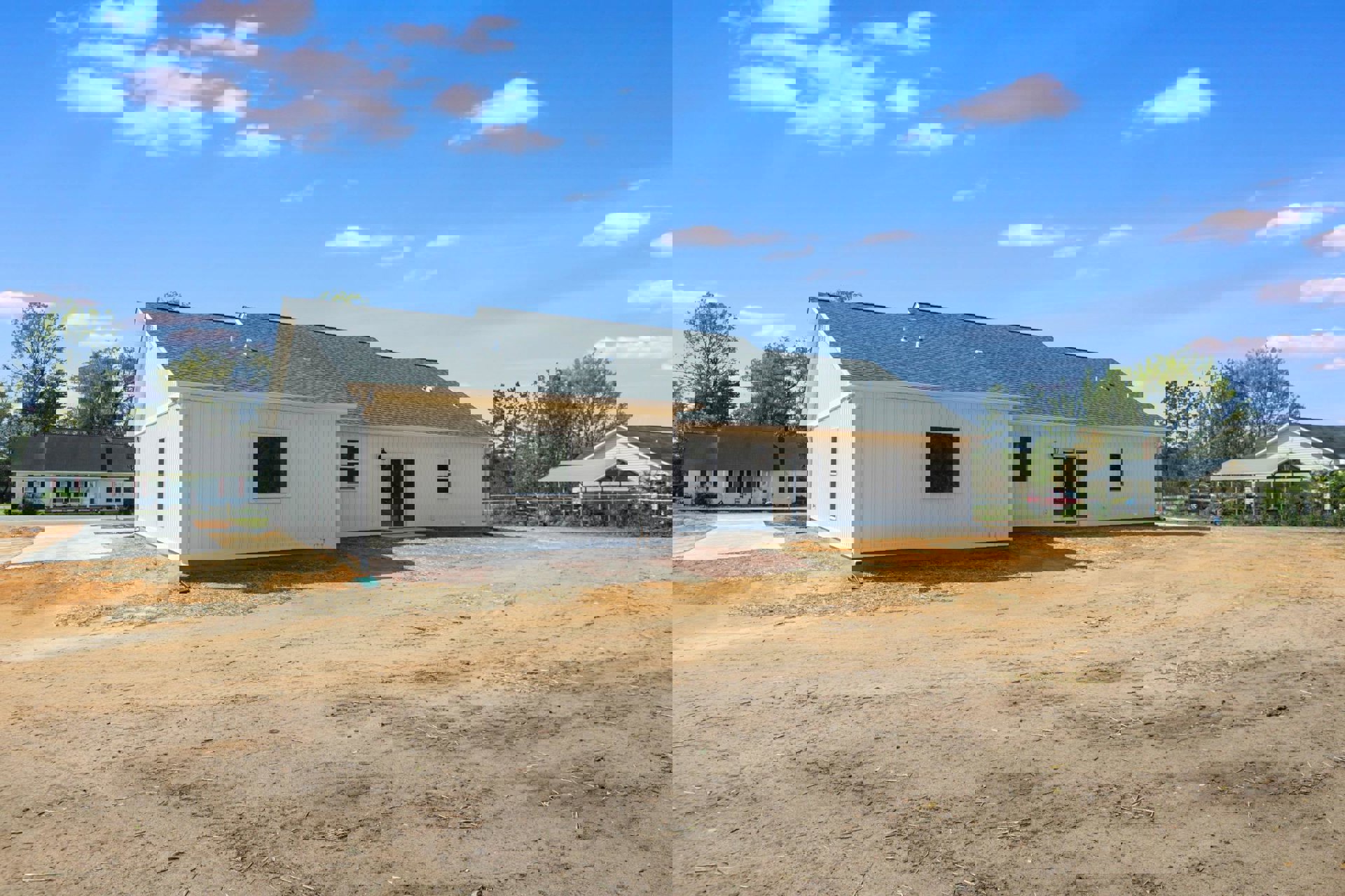 White house with black roof, white-framed windows and door, situated on a dirt lot with trees and cloudy sky in the background