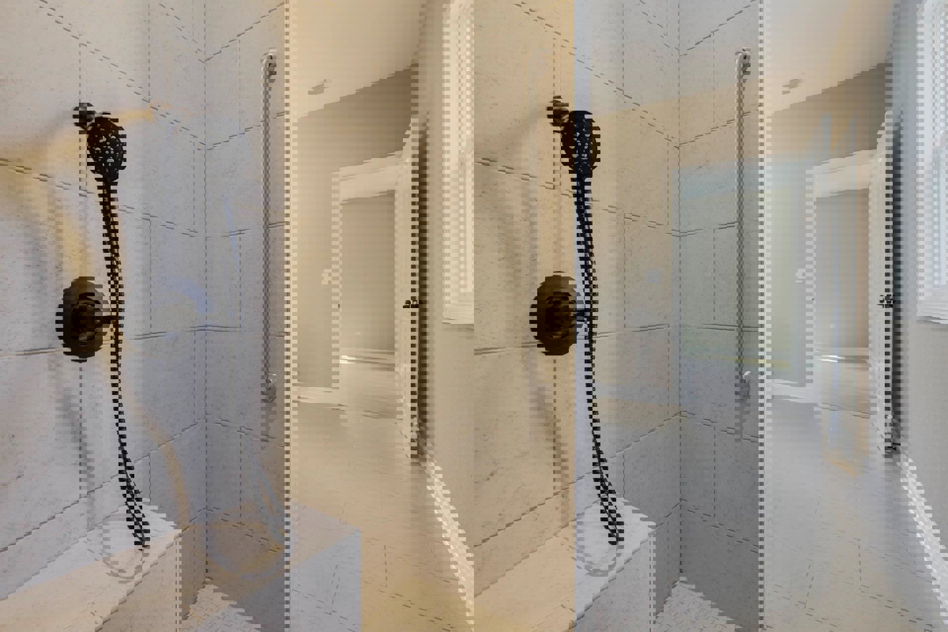 White tiled shower with a black circular hand shower mounted on a hose, black handles, and a window on the wall.