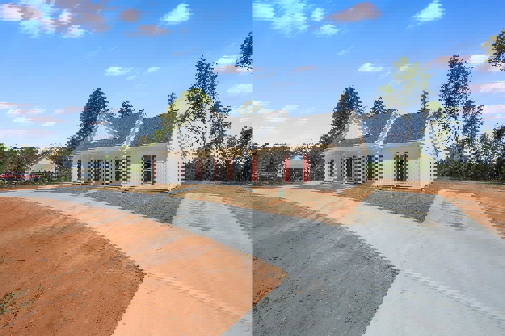 White siding house with red trim and shutters, paved driveway, white framed windows, wooden fence, mature trees in background under partly cloudy sky