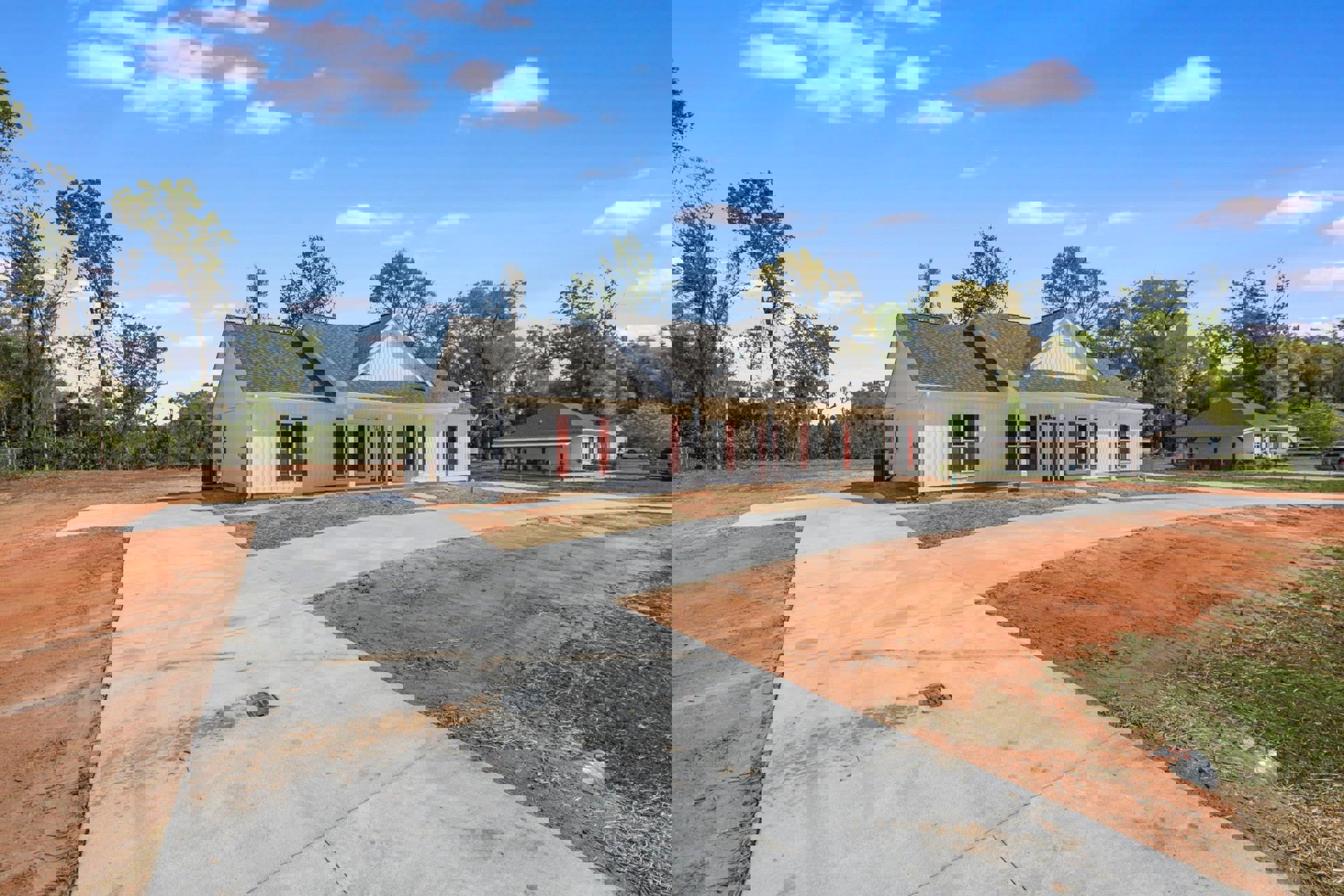 White house with red shutters, gable roof, and paved driveway bordered by dirt, surrounded by trees under a partly cloudy sky.