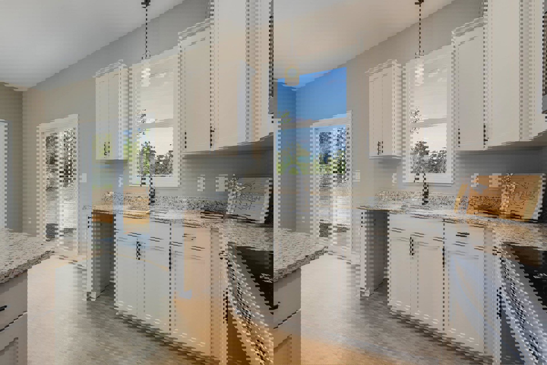 White kitchen cabinets with brushed metal handles, undermount sink beneath a window, light gray countertop, wooden cutting board with ribbon, and pillar sconce.