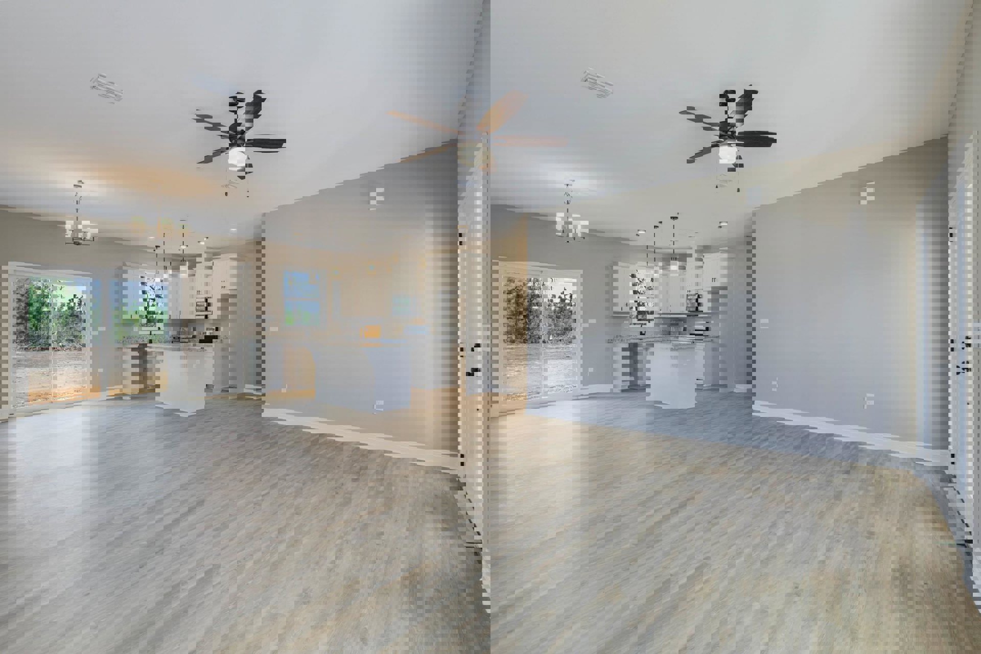 Open-concept kitchen and dining area with wood flooring, white cabinetry, marble countertops, ceiling fan with light, and window overlooking a dirt road and trees.