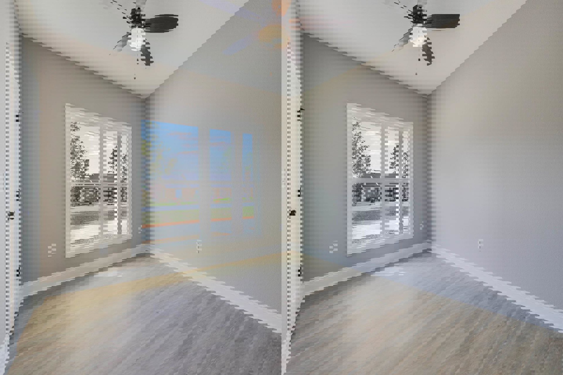 Bright room with white wood flooring, white walls and trim, ceiling fan with light, large window overlooking street and houses, white door with black handle