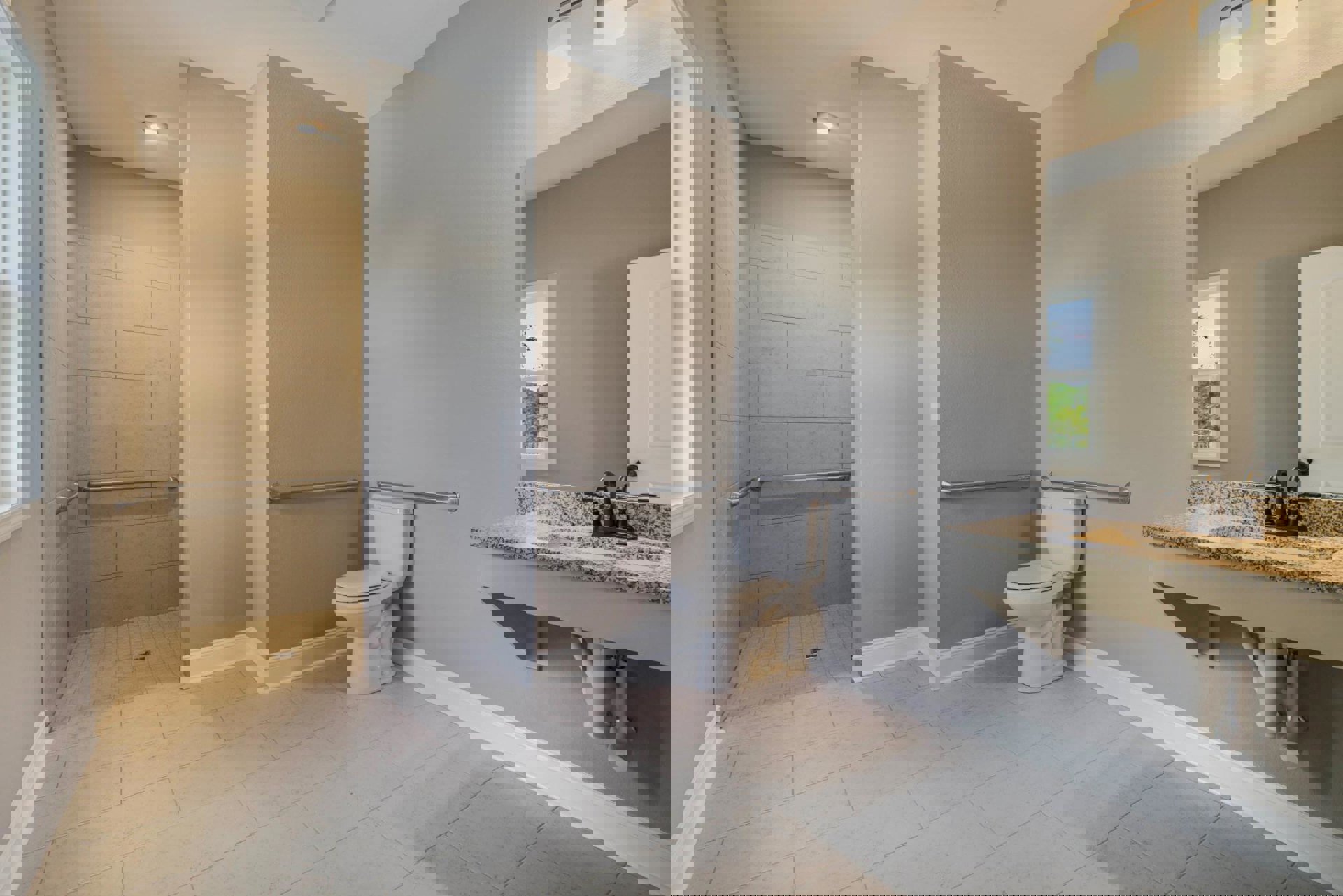 White porcelain sink and toilet on light tile floor, large window with tree view, wall-mounted mirror above stone countertop, white door partially visible.