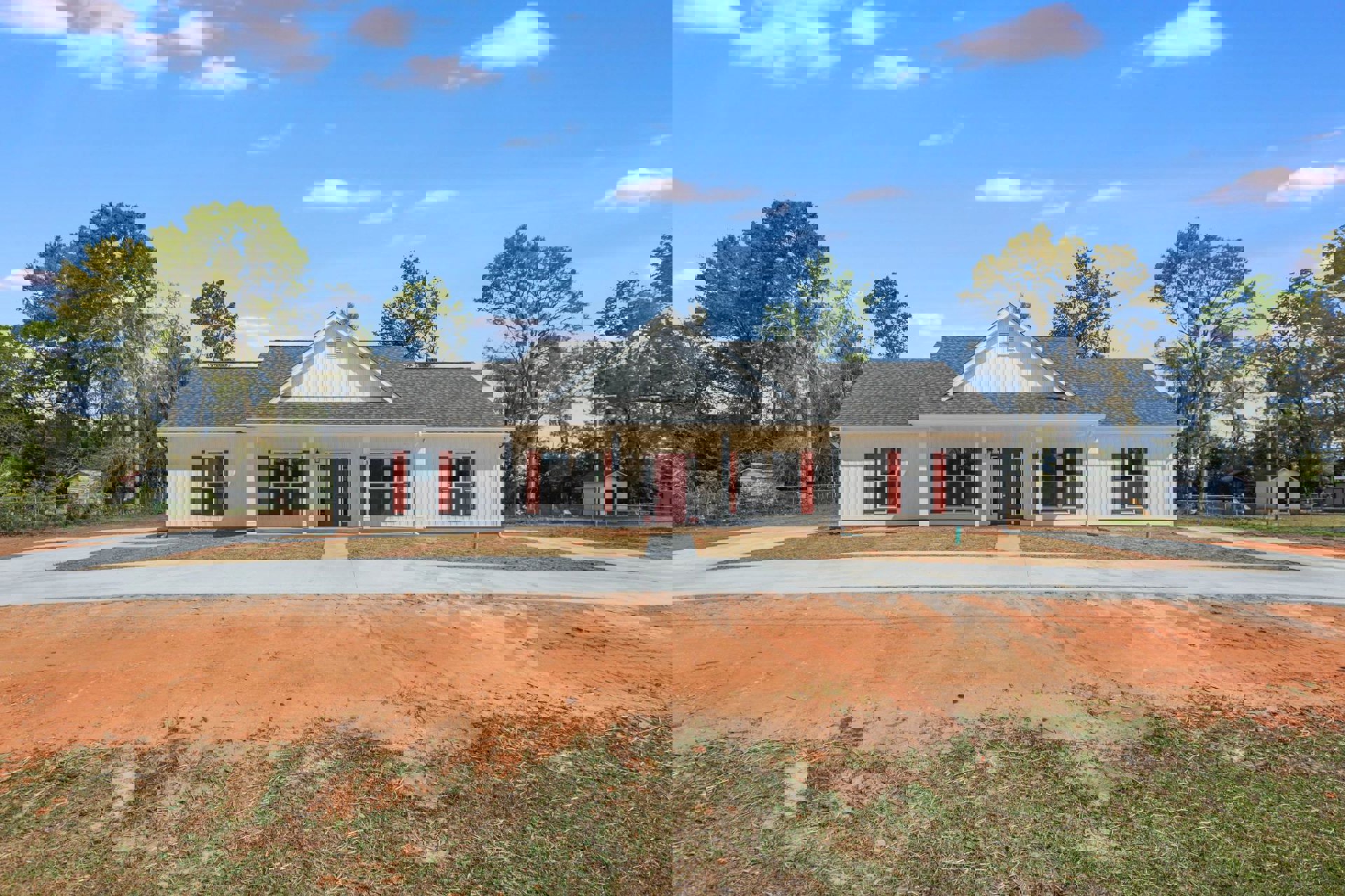White siding house with red shutters and red front door, tree in front yard, dirt road bordered by grass, windows with glass panes, roof and trees in background under cloudy sky