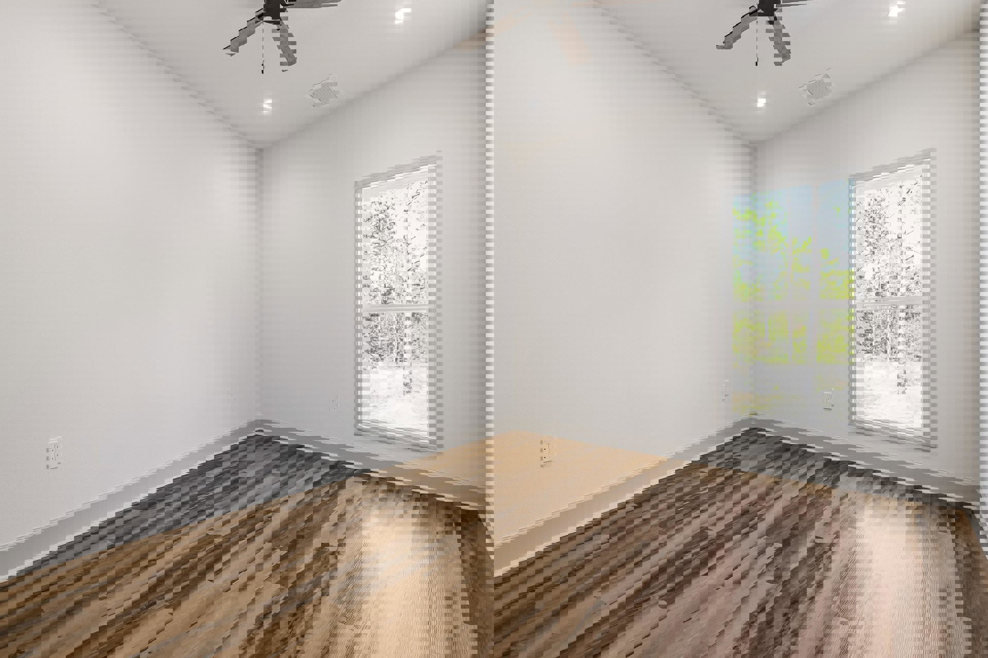 Ceiling fan with light fixture mounted on white plaster ceiling, wood flooring throughout room, large window with view of trees, wall vent near ceiling