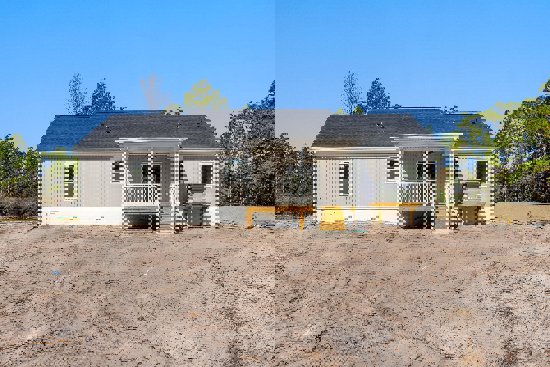 Two-story house with white siding, elevated wooden deck and stairs leading to sandy dirt yard, white door with window, surrounded by trees under blue sky