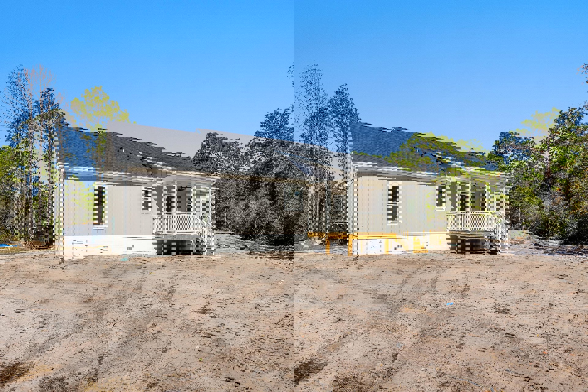 Two-story house with white-framed windows, sandy dirt yard, wooden deck, covered porch, black dumpster, and blue sky overhead