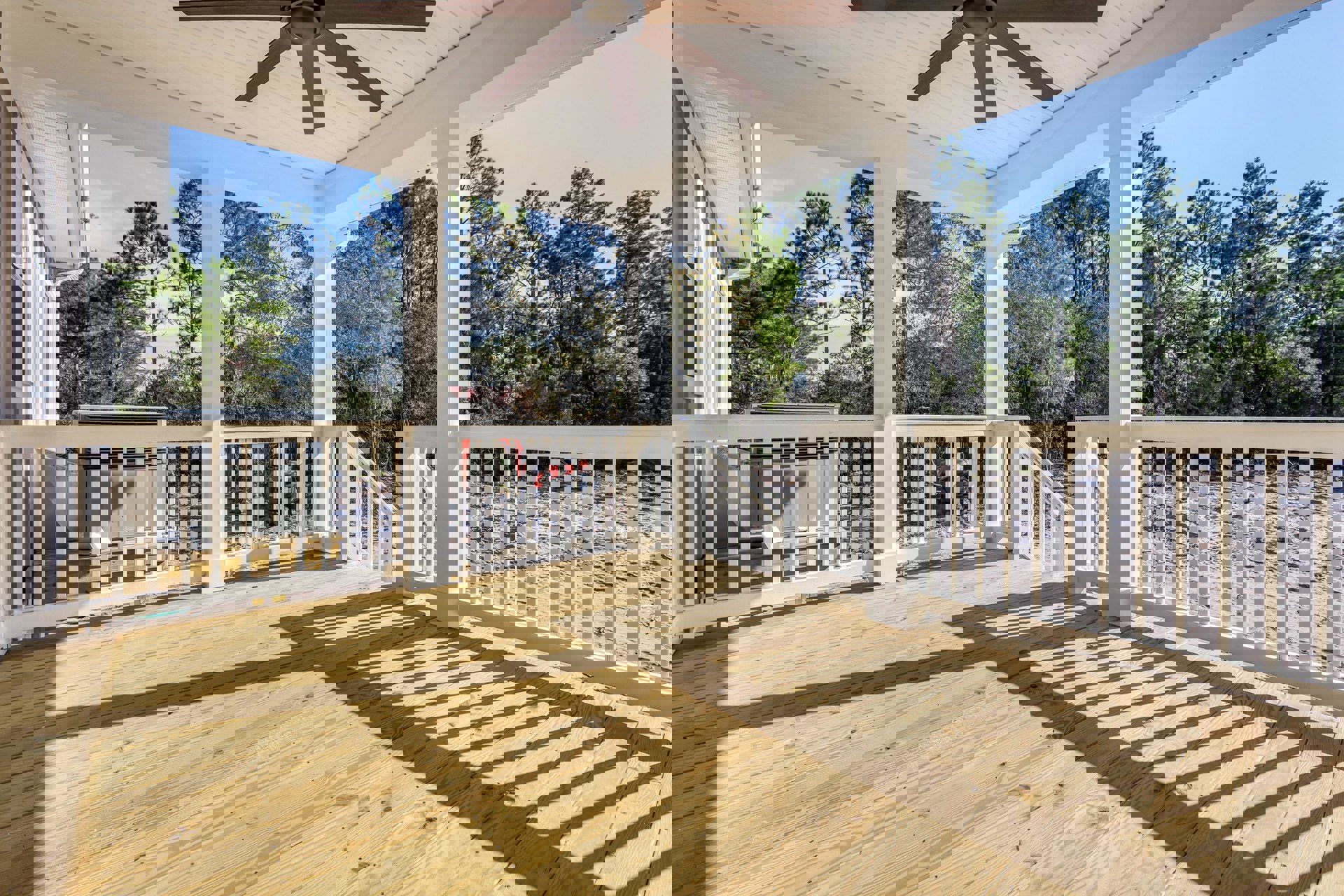 Wooden deck porch with white railings and pillars, ceiling fan overhead, surrounded by trees under a blue sky