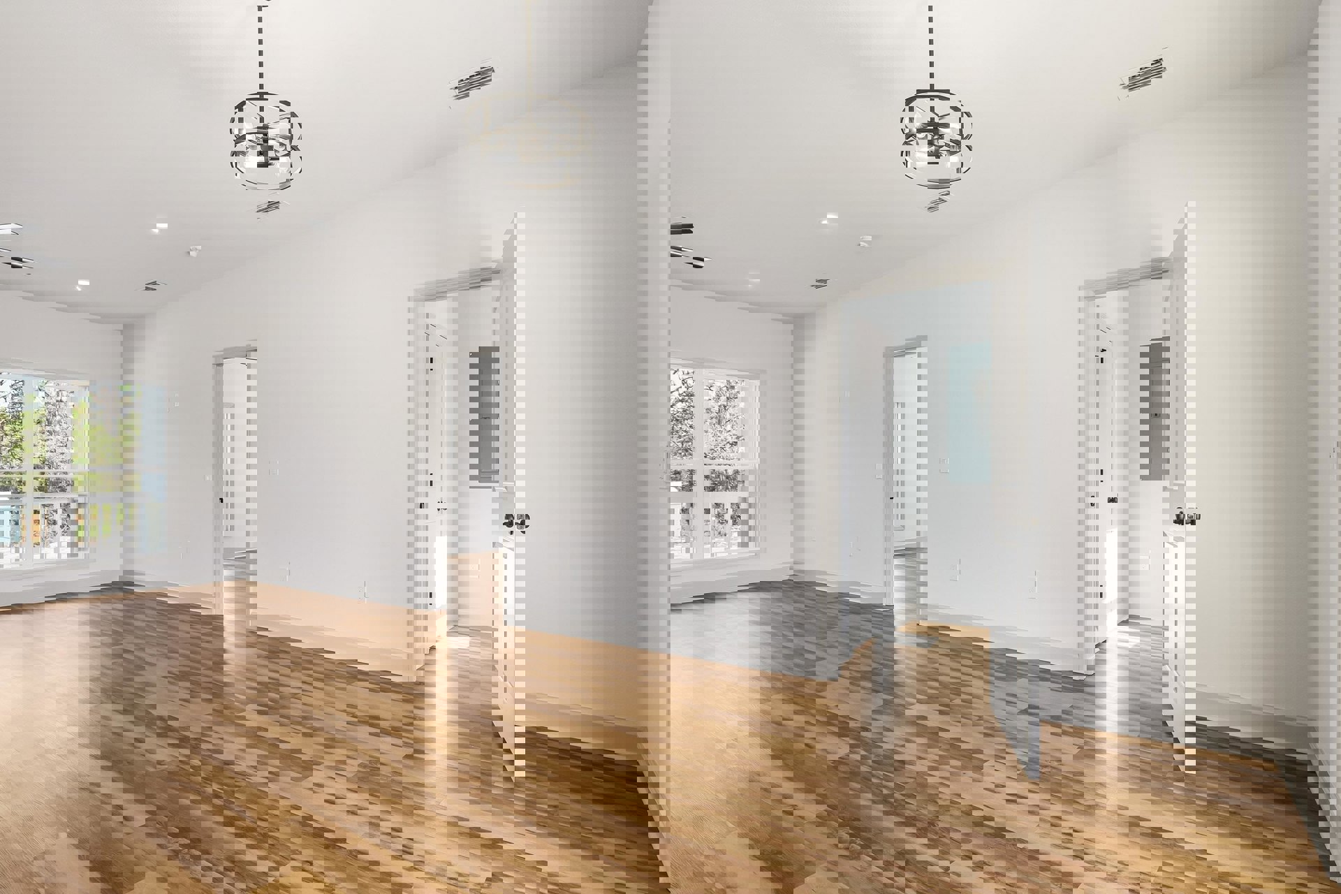 Hardwood floor and white walls in a bright room, metal-framed light fixture overhead, window with white railing, wall vent, and grey door with a square control panel.