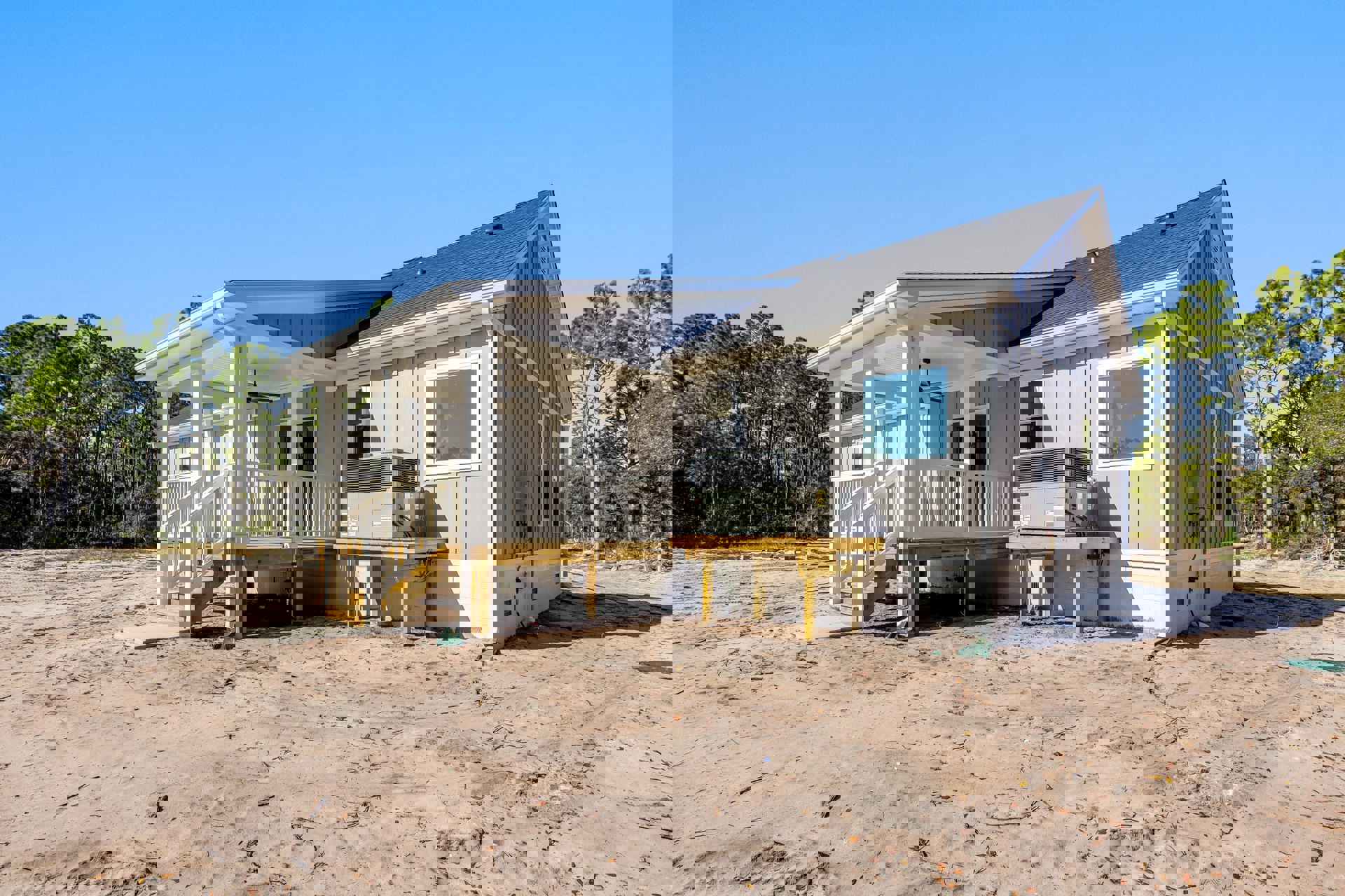 Grey house with white porch, large windows, and air conditioner unit, set on a dirt field with sparse trees in the background.