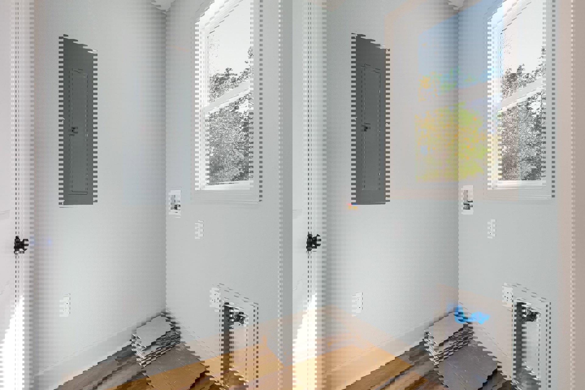 Wood plank flooring in a room with a large window overlooking trees, grey door with square cutout, stacked tiles, close-up wall outlet, blue plastic bag in metal bin, white plaster