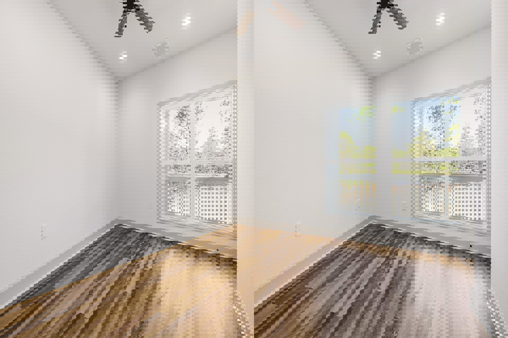 Sunlit room with large window overlooking trees, wood flooring, white plaster walls, ceiling fan, and ceiling vent