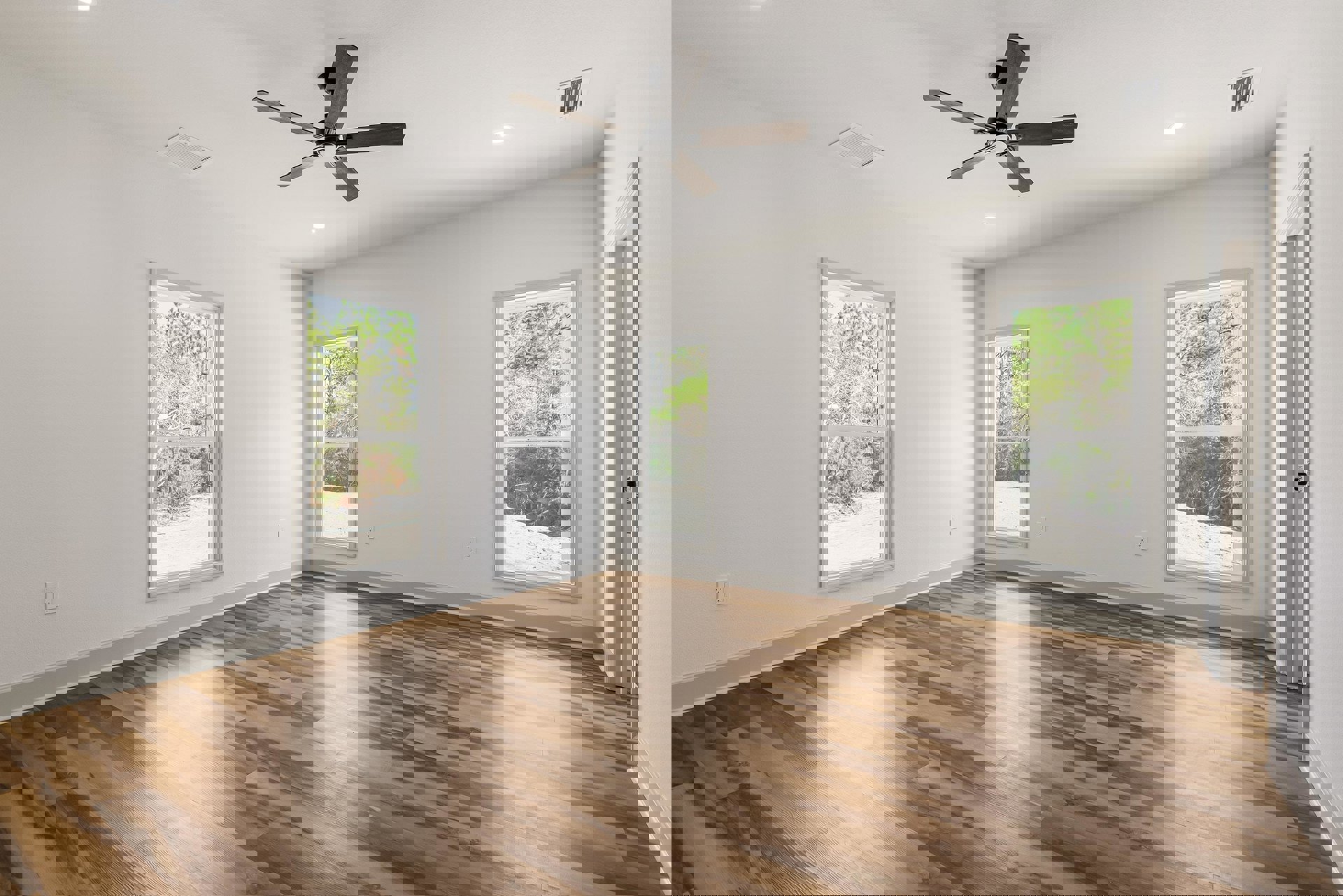 Ceiling fan with light fixture, wood flooring, white-framed windows showing trees outside, plaster walls and ceiling, floor vent beneath window