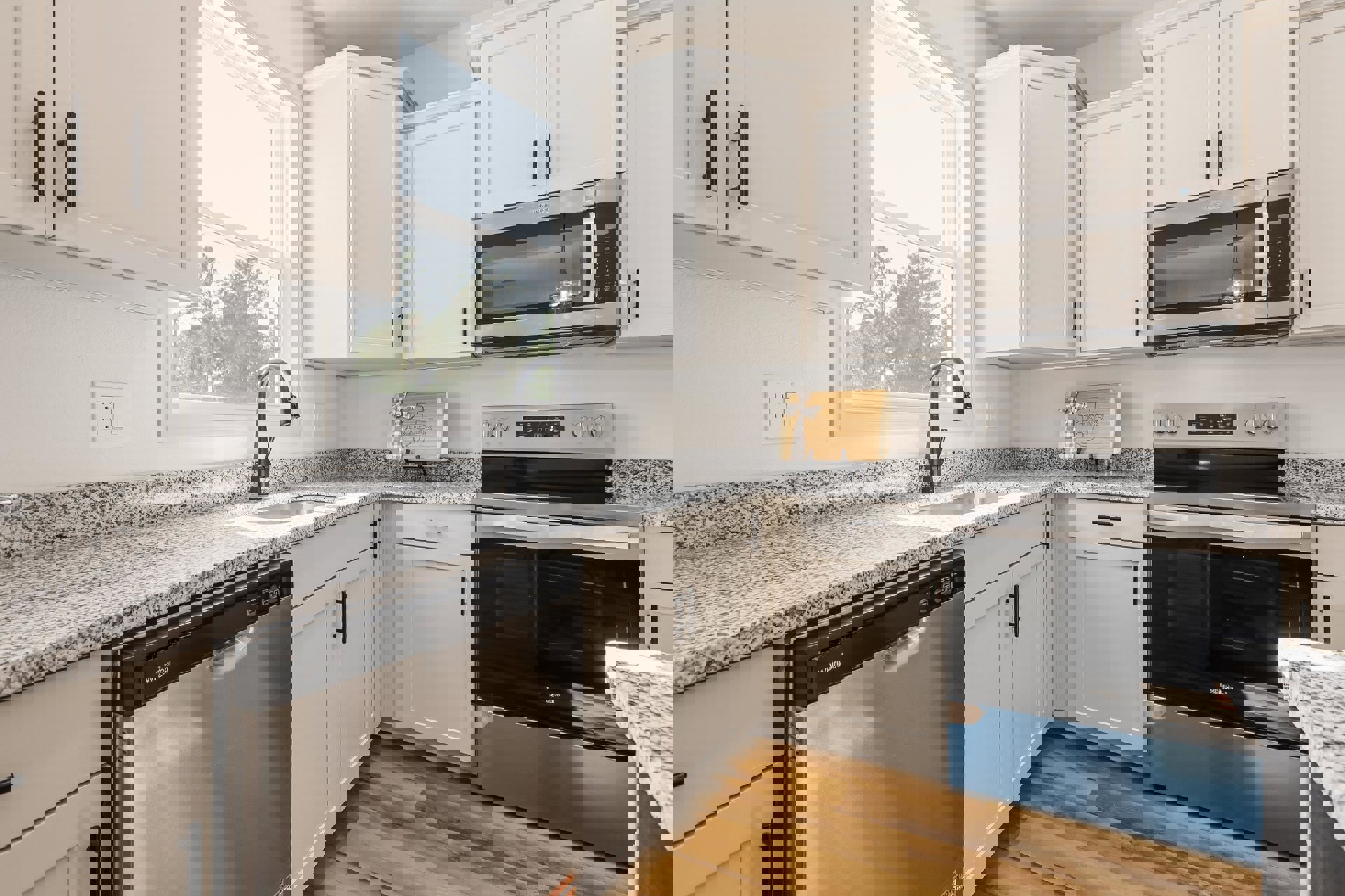 White shaker cabinets, black dishwasher under quartz countertop, stainless steel oven and microwave, black faucet, wooden cutting board with ribbon on counter