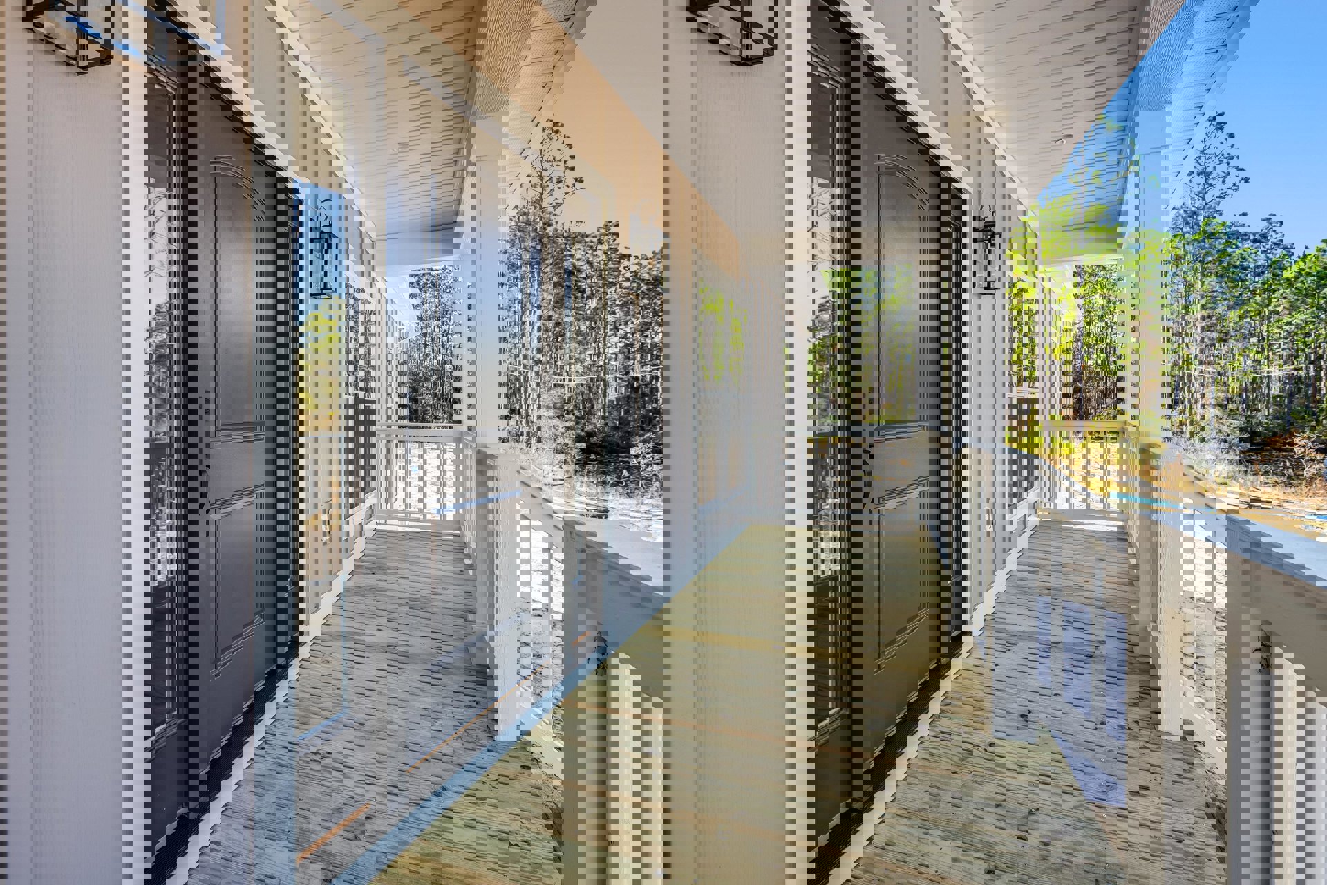 Front porch with white ceiling and light fixture, black door with handle, glass staircase railing, deck featuring white railing, surrounded by trees and sky.