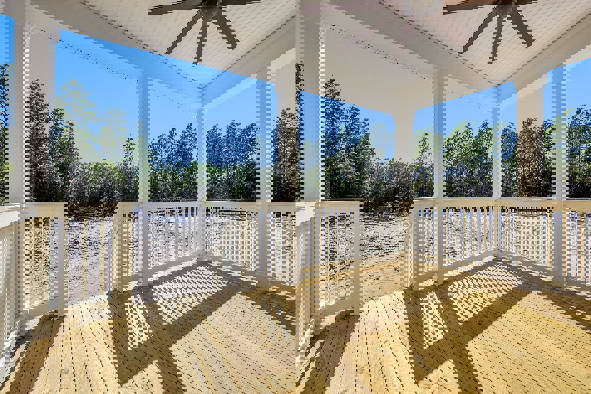 Wooden deck with white railings, ceiling fan overhead, and leafy trees in the background
