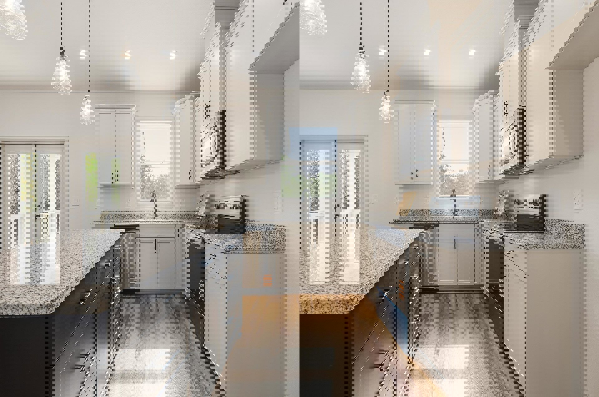 White kitchen cabinets and granite countertops, stainless steel microwave and stove, wooden cutting board with a bow, pendant light fixture, window overlooking trees and blue sky