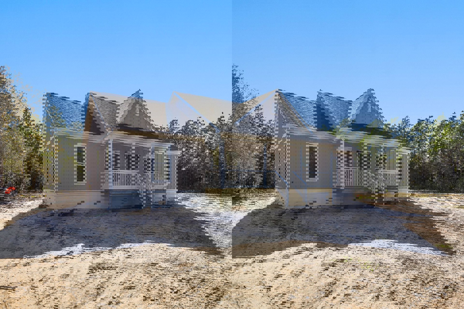 Two-story house under construction with white-framed windows, covered porch, sandy dirt yard, white railing, and visible roof; tractor parked near unfinished stairs, trees in
