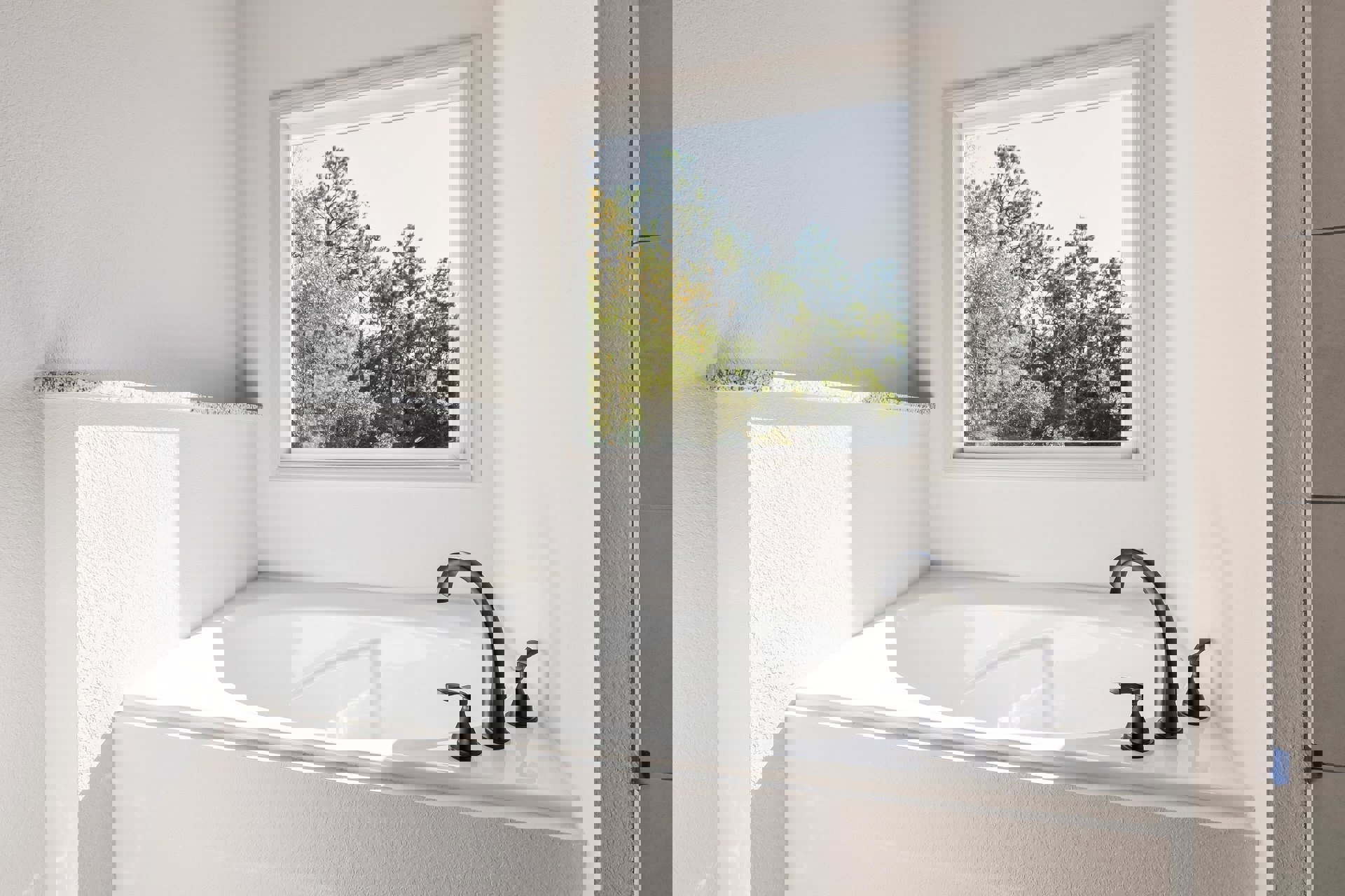 Freestanding white bathtub with matte black faucet beneath a window overlooking trees, surrounded by light tile walls