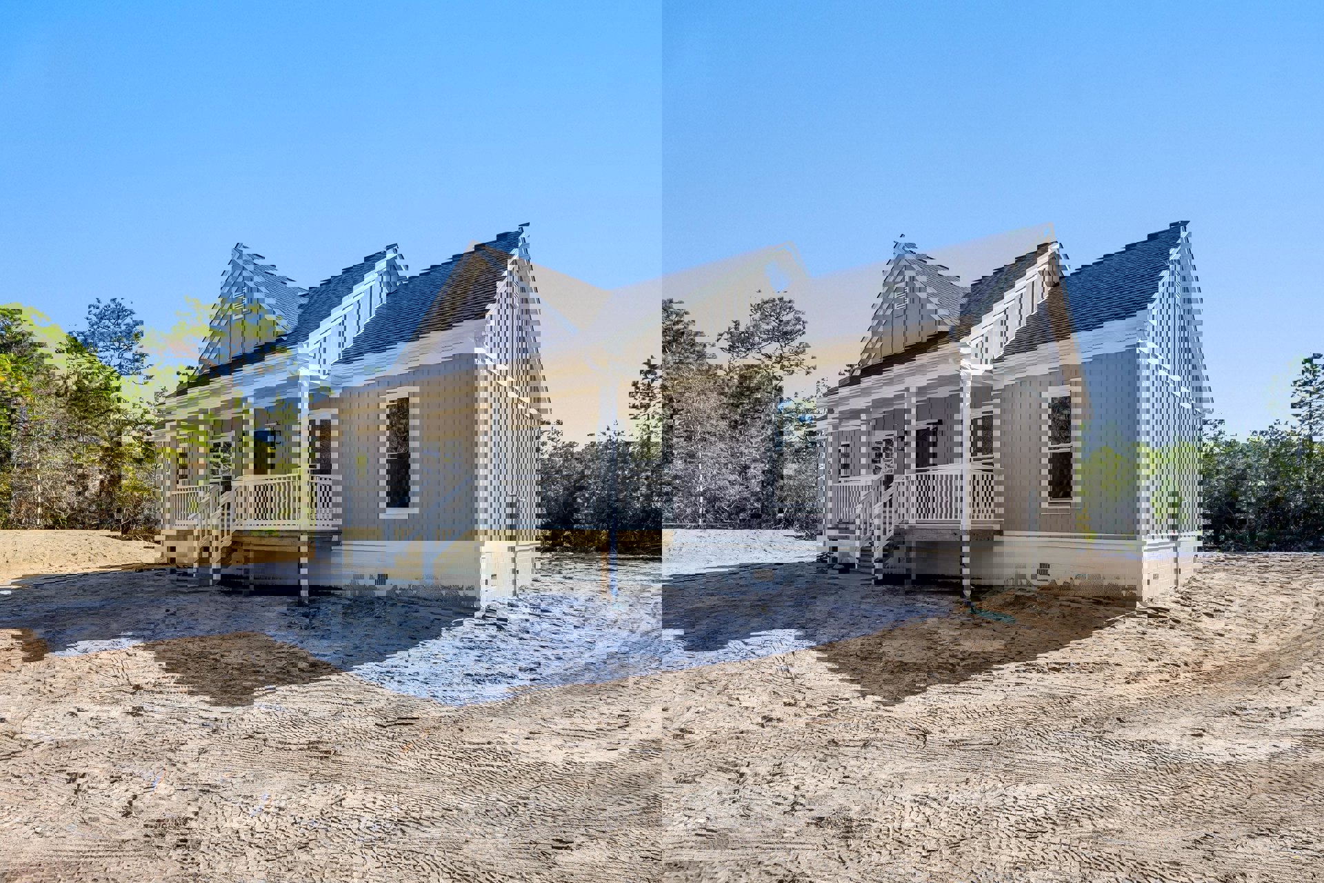 Partially built house with white-framed windows, wooden porch featuring white railing, sandy ground marked by tire tracks, and a leafy green tree nearby