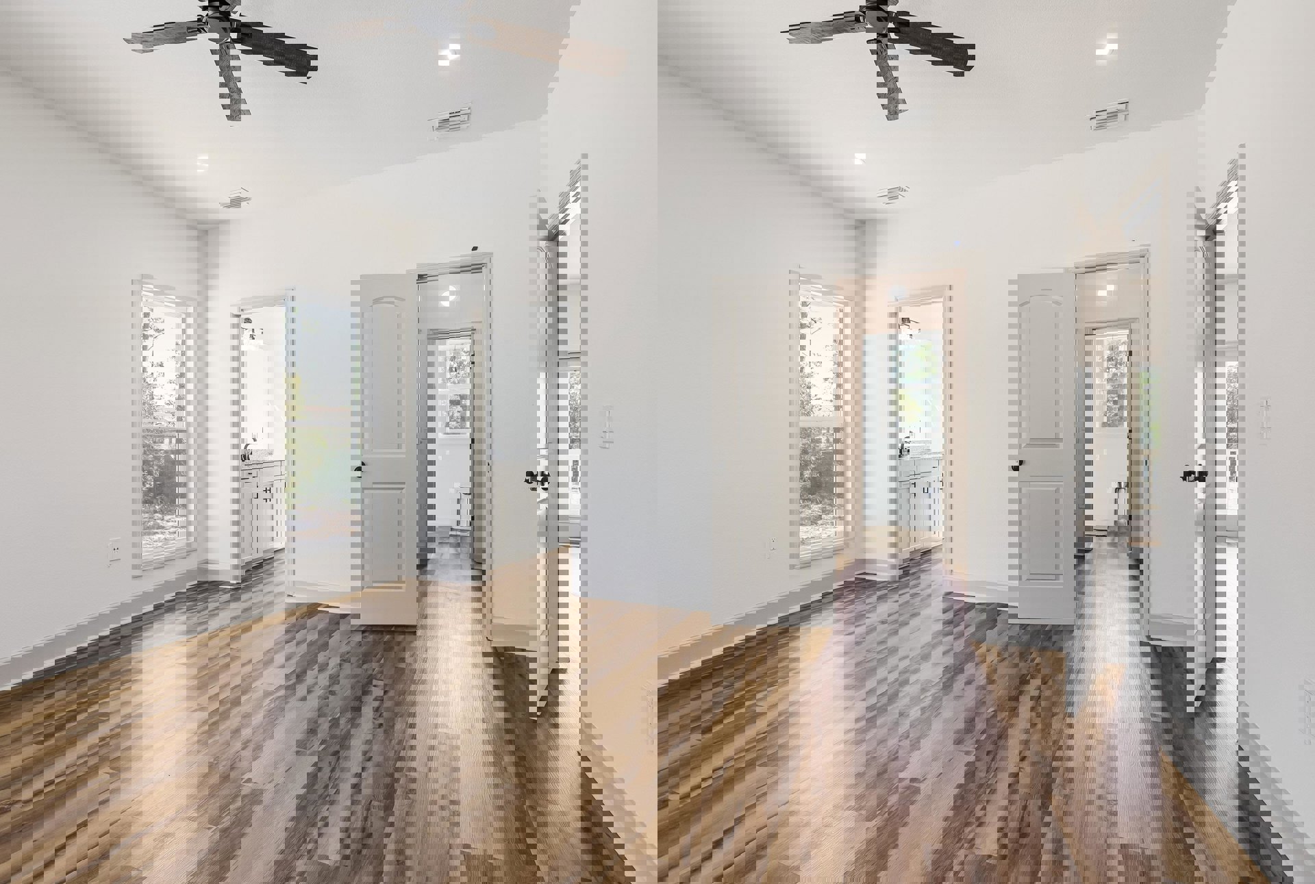 White walls, wood flooring, window overlooking trees, white door with black knob, ceiling fan, bathroom with sink and mirror