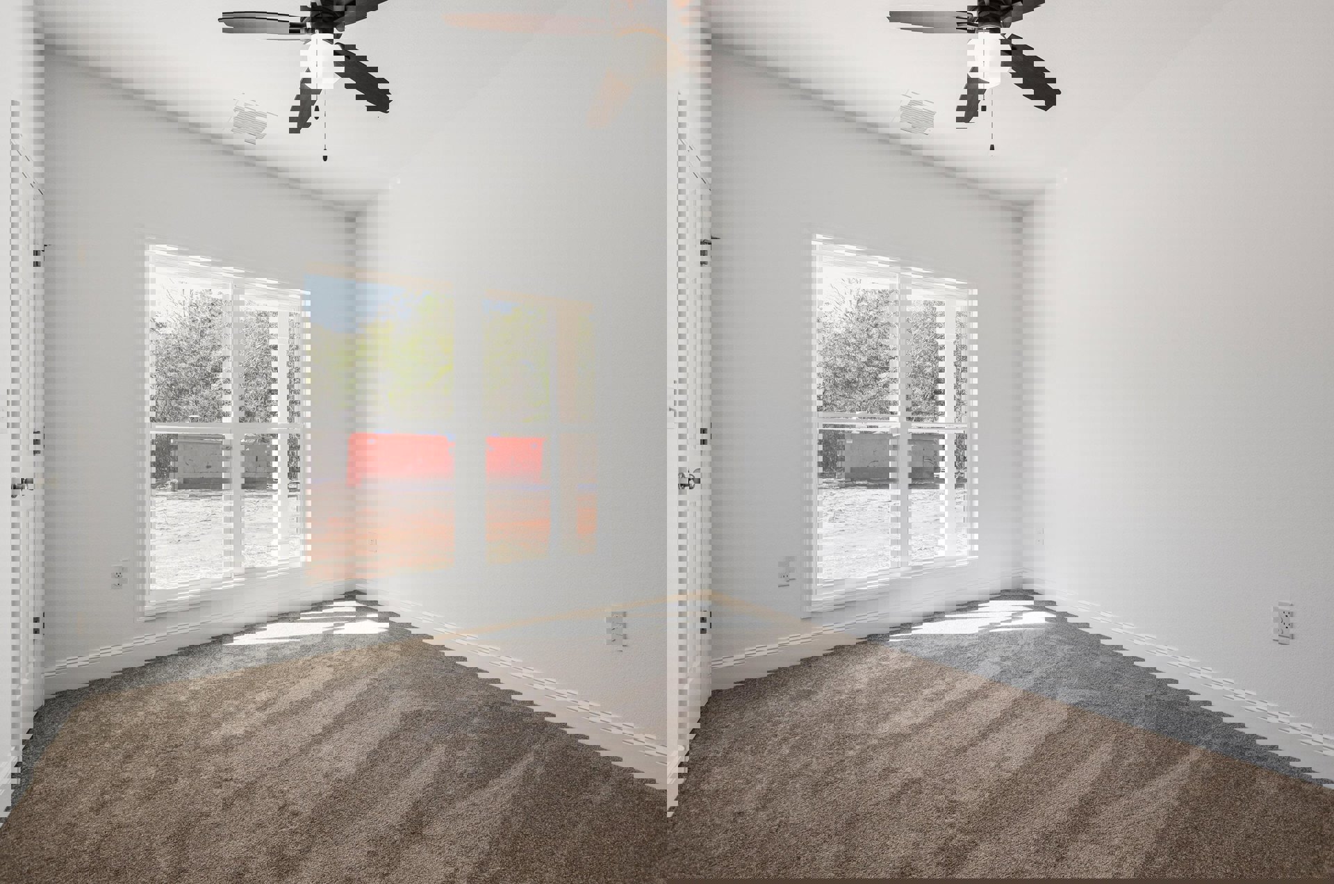 Bedroom with light wood flooring, white plaster walls, ceiling fan with integrated light, large window offering view of red shipping container, and ceiling air vent.