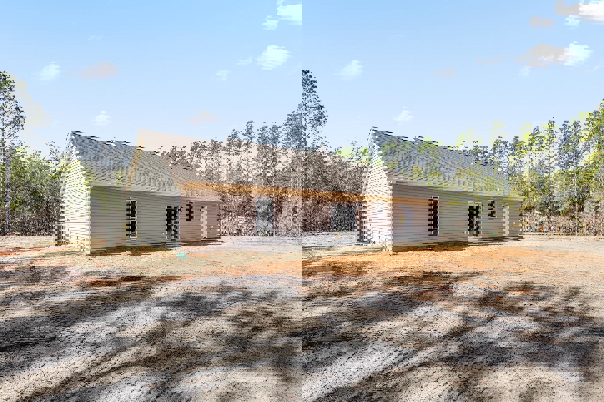 Single-story house with beige siding, several windows including one with broken glass, dirt yard in front, dirt road leading to property, large tree with many branches, shed