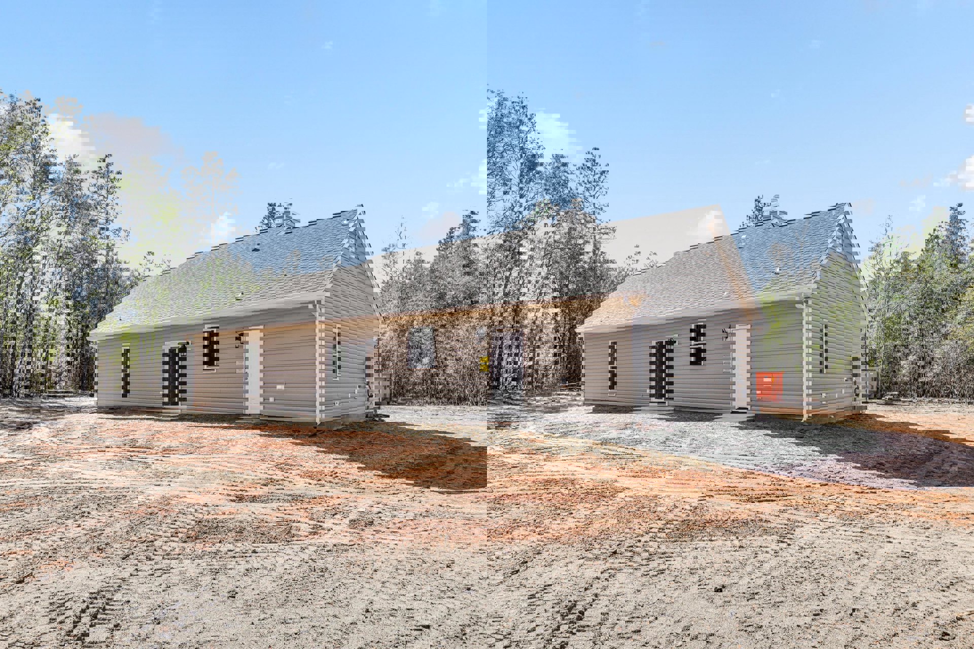 Partially built house surrounded by dirt and mature trees, white front door installed, window visible on facade, construction materials and containers scattered on ground.