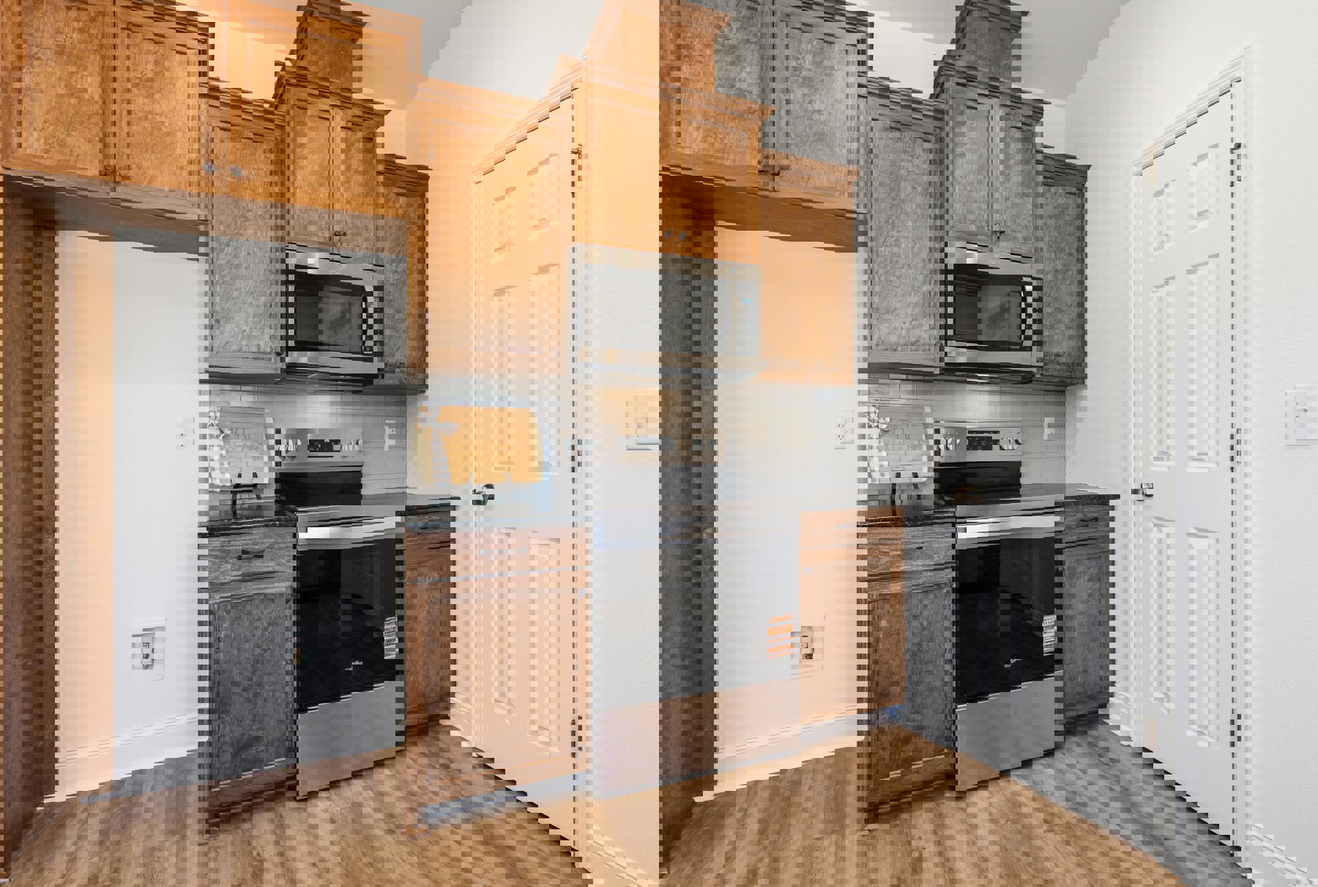 Kitchen with natural wood cabinets, stainless steel microwave and oven, black countertop, wooden cutting board, and white door with silver knob