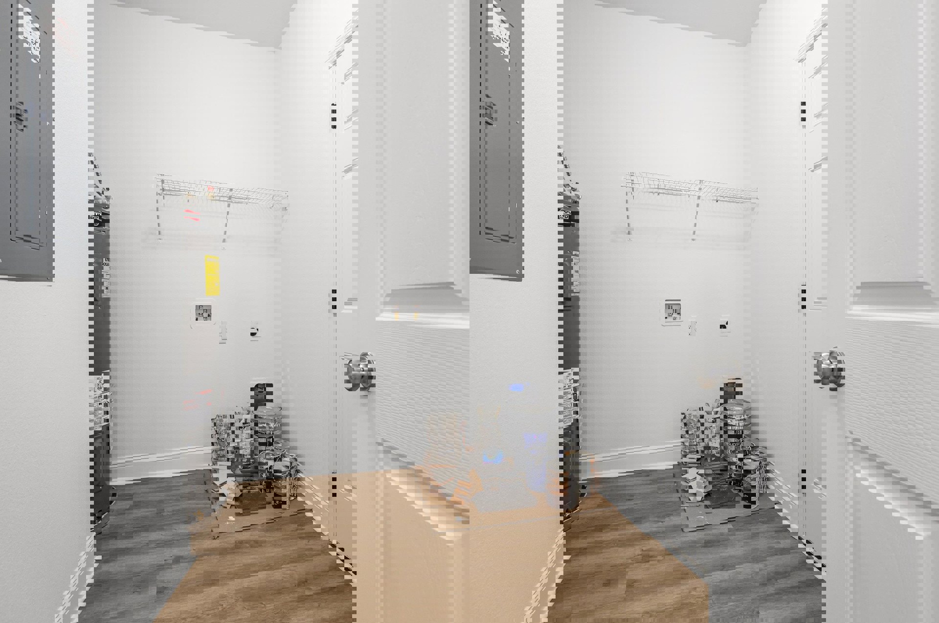 Open door leading to a room with white walls, wood flooring, cardboard boxes, and cabinetry; visible bathroom sink and tap fixtures.