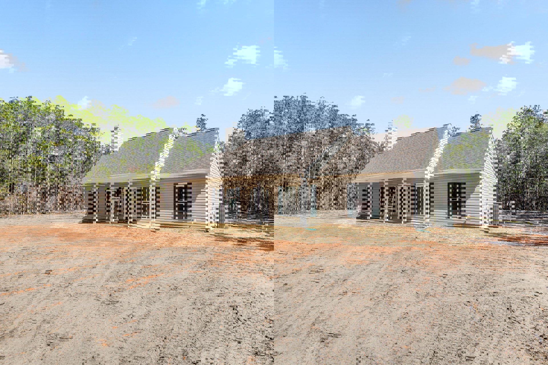 Two-story house with white-framed windows, covered front porch, gray roof with chimney, surrounded by dirt yard and scattered trees under a cloudy sky