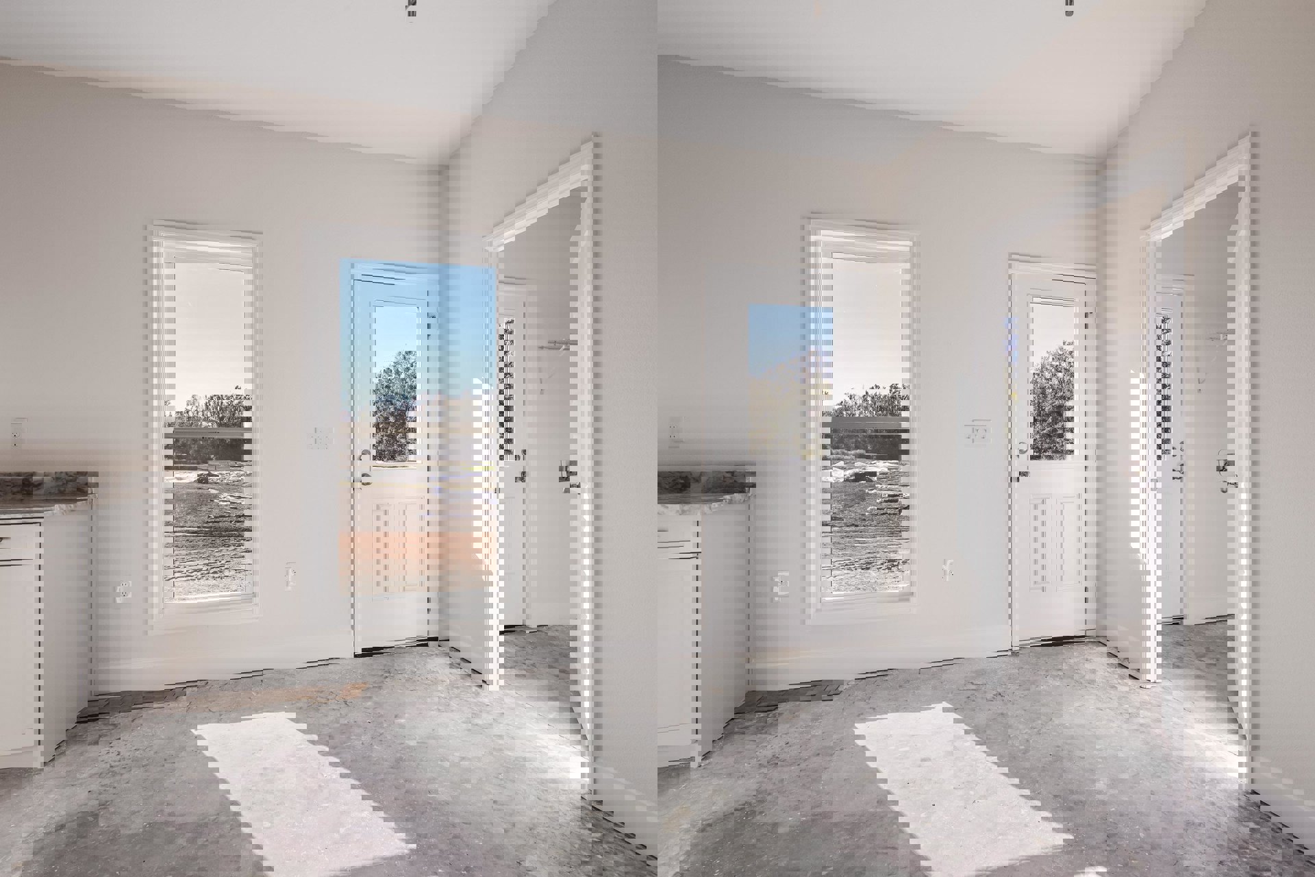 White-walled room with two doors, concrete floor, large windows showing a field and trees, white cabinet with drawer, sunlight casting shadows on white surfaces